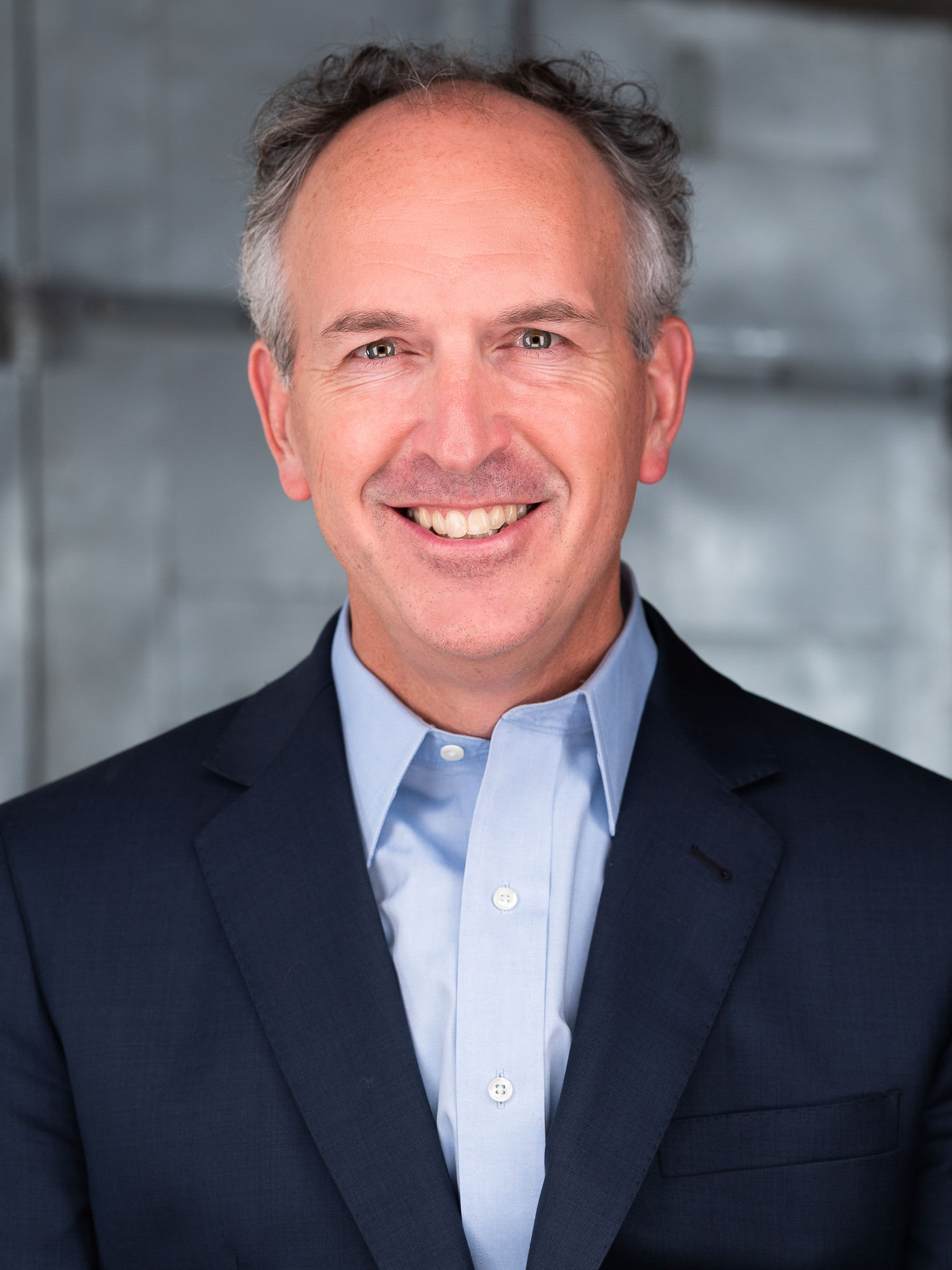 A professional headshot of a man wearing a blue button down and suit jacket. He has short gray hair and he is smiling looking straight ahead.