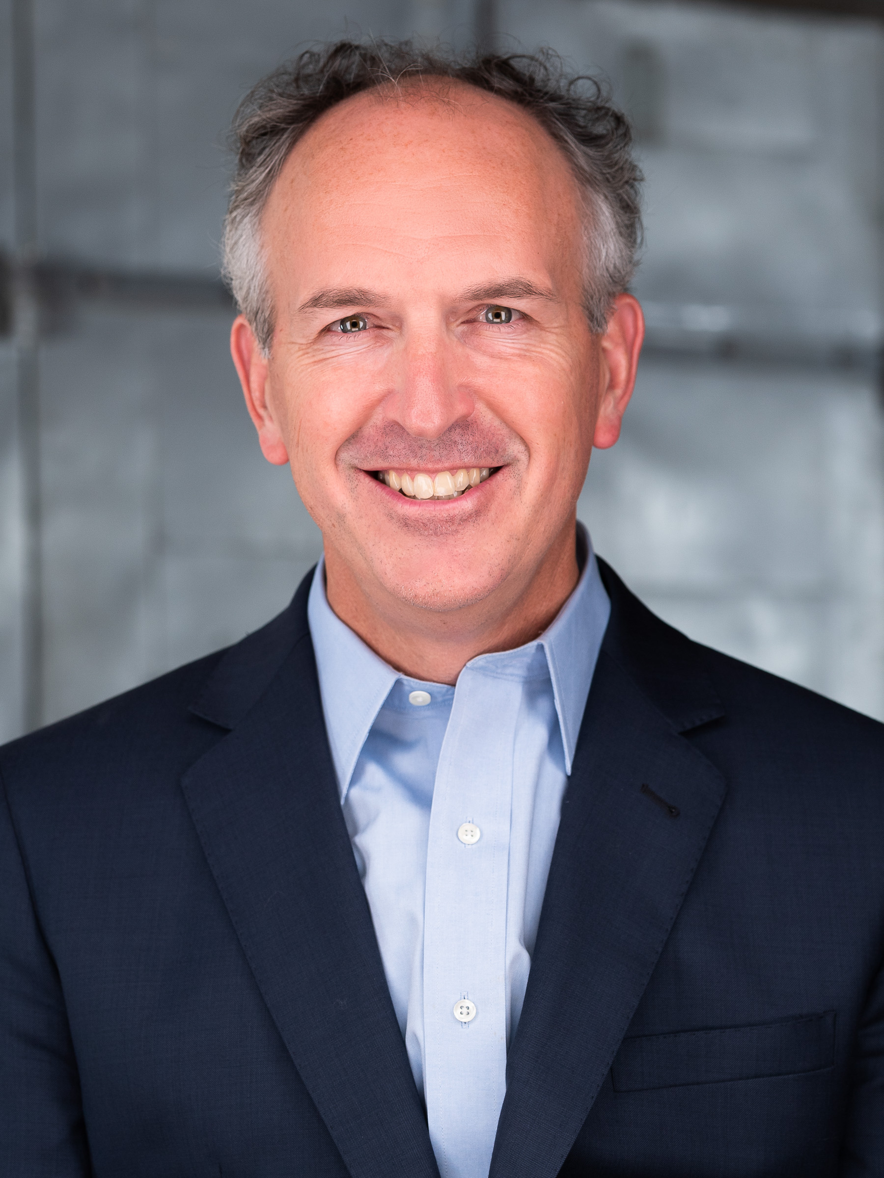 A professional headshot of a man wearing a blue button down and suit jacket. He has short gray hair and he is smiling looking straight ahead.