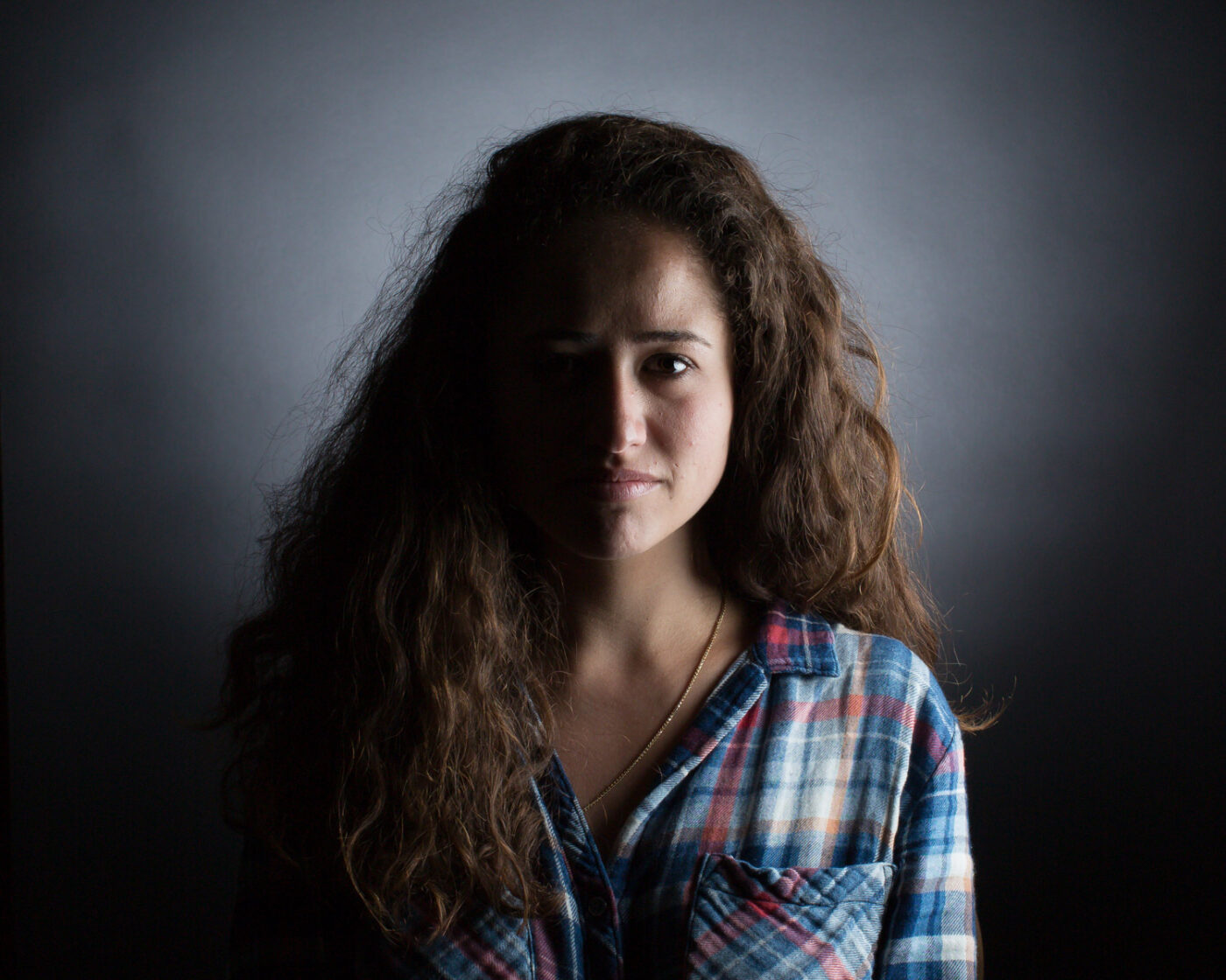 A portrait from the Shine a Light on Usher Syndrome Exhibit featuring a young woman with tan skin and brown curly hair. The woman's face is half in shadow and half in light and she wears a plaid button down shirt. The background is a gray gradient.