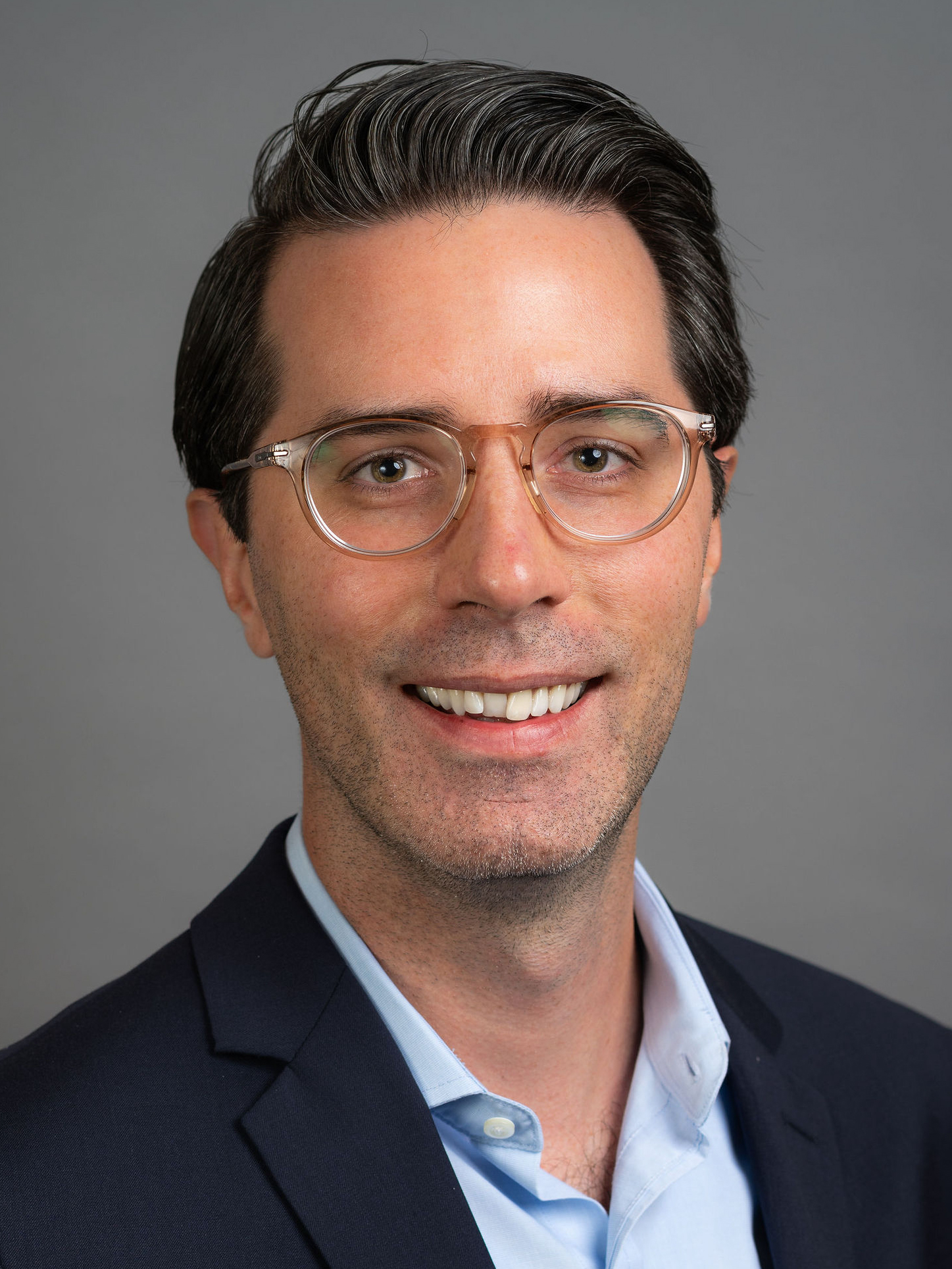 Close up headshot of Eliot Shearer. He wears a blue collared shirt and dark jacket. He has short brown hair and glasses.