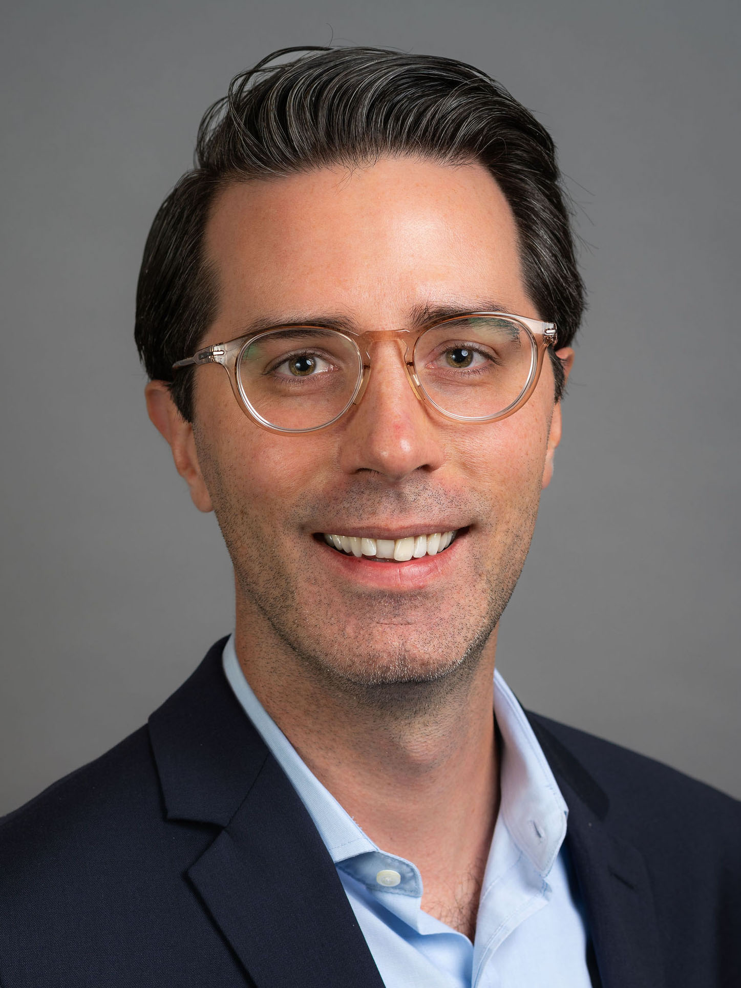 Close up headshot of Eliot Shearer. He wears a blue collared shirt and dark jacket. He has short brown hair and glasses.