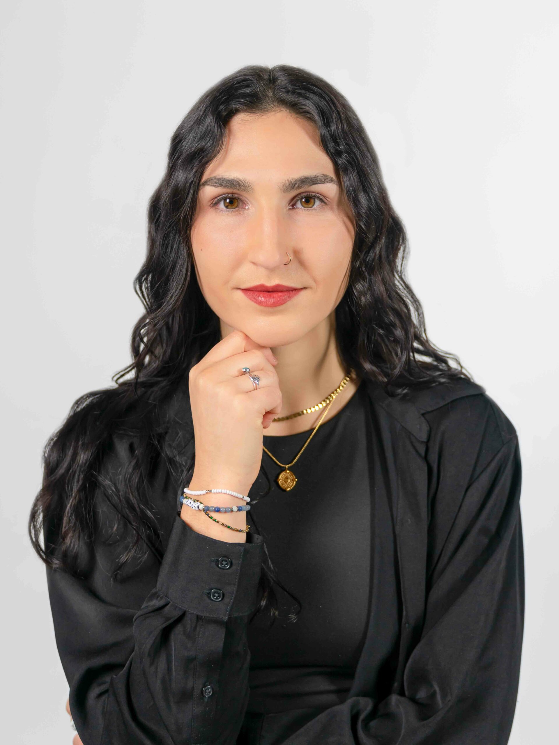 A headshot of a young woman, Eliza Corderman, standing with one hand to her chin. She has wavy black hair and wears a black shirt. She looks out straight ahead.