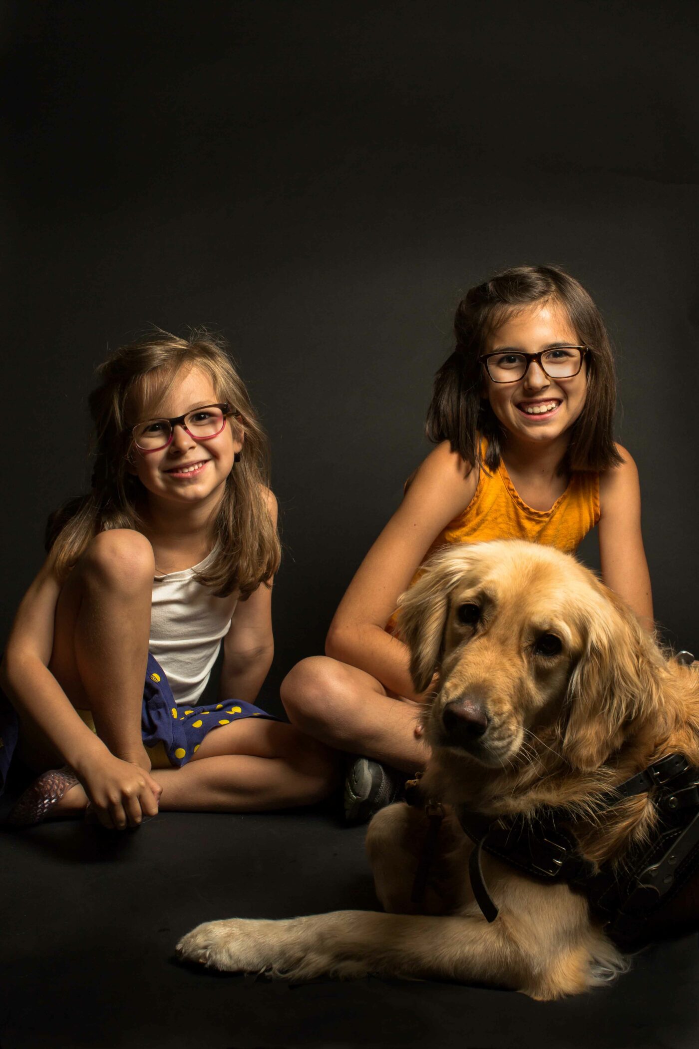 A portrait from the Shine a Light on Usher Syndrome Exhibit featuring two girls seated on the floor next to each other and with a golden retriever dog. Both girls look ahead and are smiling with wide smiles. One of the girls is older than the other girl. The younger girl is seated with one of her knees pulled up into her chest and she wraps one arm around her leg. She has medium-length light brown hair and she wears glasses that have a pink frame on the bottom of the front lens. She wears a white shirt and blue skirt with yellow polka dots. The older girl is seated with her legs crossed right behind the golden retriever. She has shoulder length brown hair and she wears glasses that have brown frames. She wears a bright orange shirt. The dog looks ahead and he wears a black harness. There is a dark gradient background behind them.