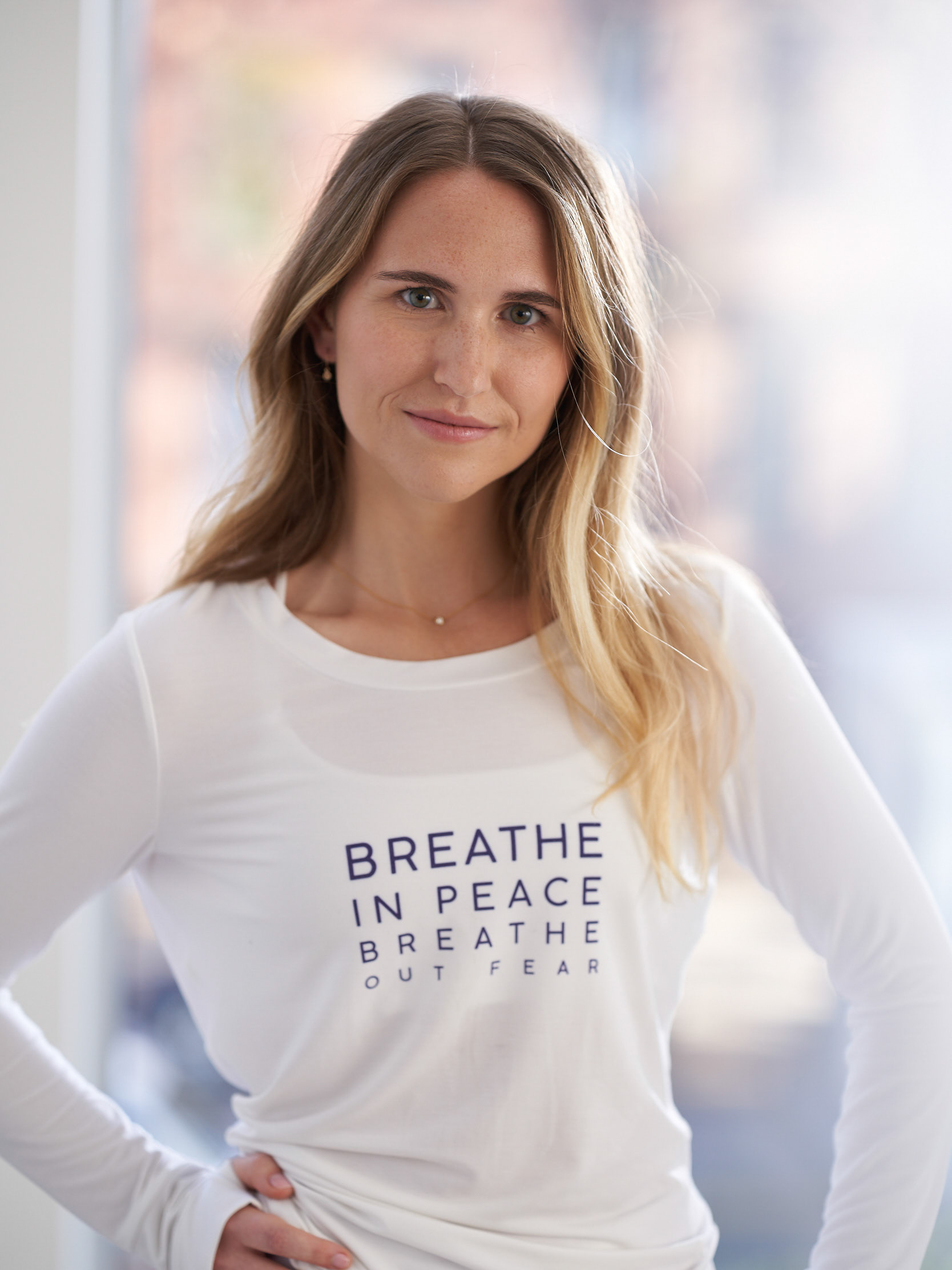 A headshot of a young woman, Hannah Corderman. She has blonde hair and stands with her hands on her hips. She wears a white long sleeve t-shirt from the Tactile collection.