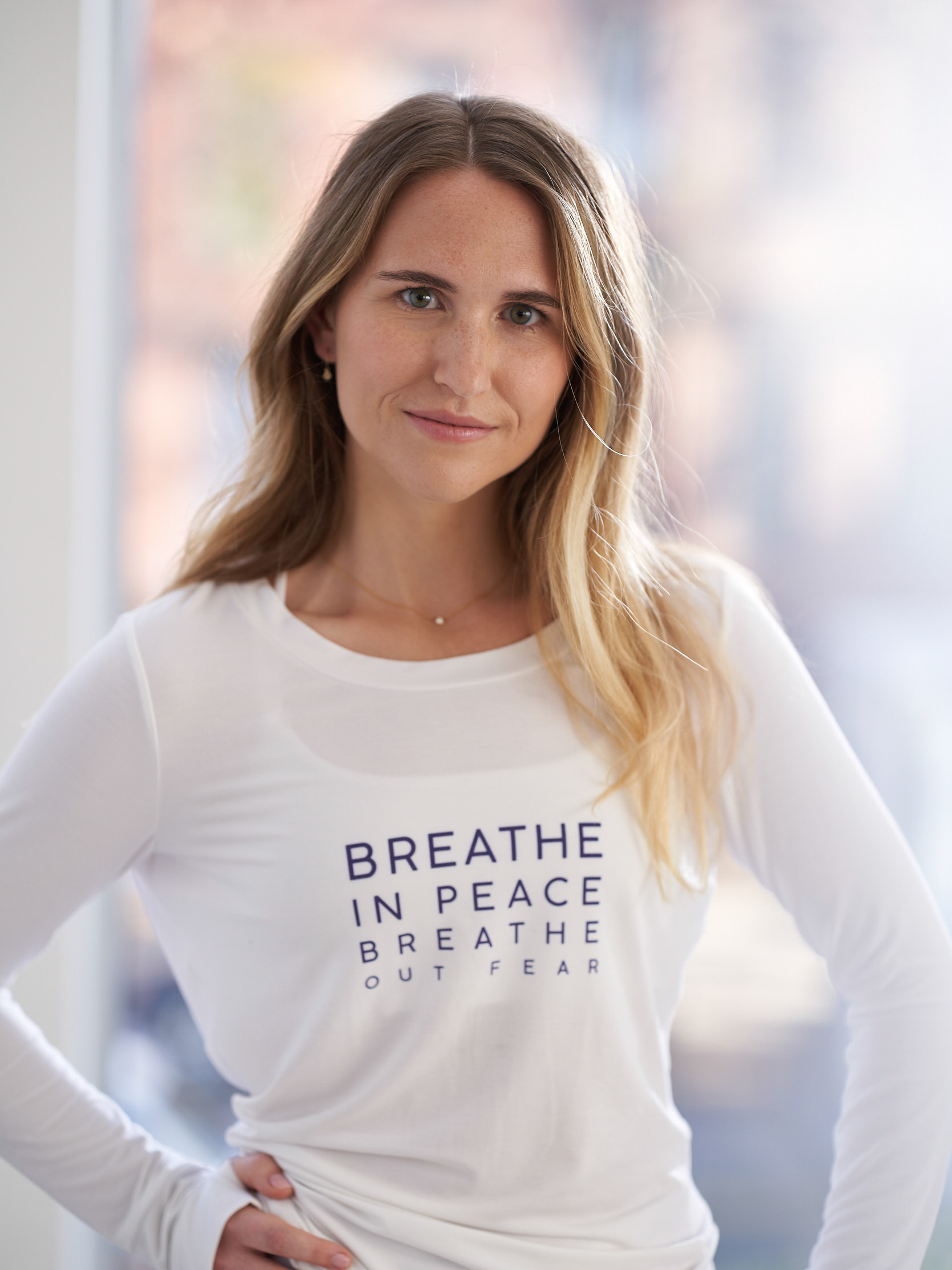 A headshot of a young woman, Hannah Corderman. She has blonde hair and stands with her hands on her hips. She wears a white long sleeve t-shirt from the Tactile collection.