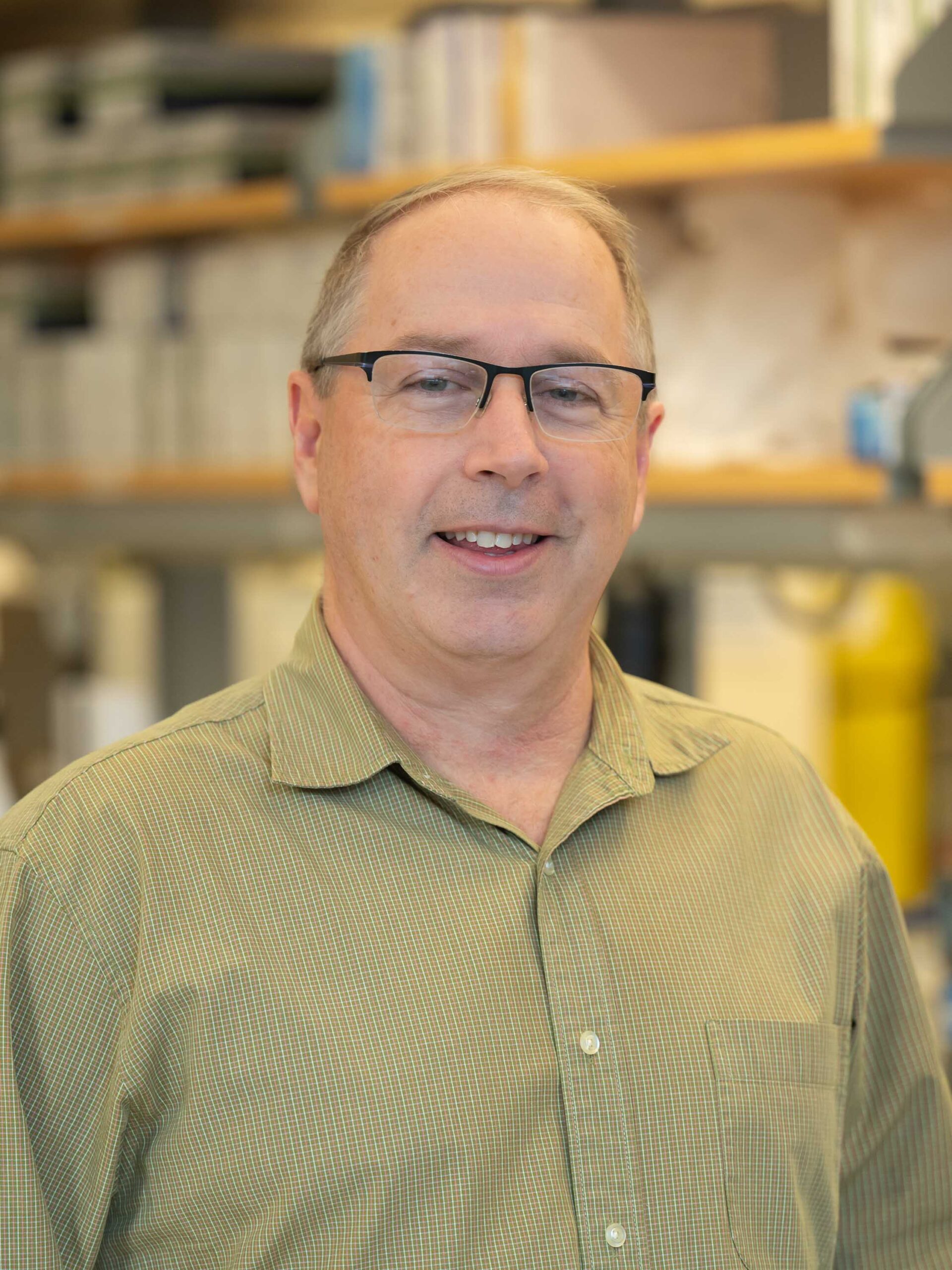 Headshot of Jeff Holt. He is standing in a lab, wearing a green button down shirt and glasses
