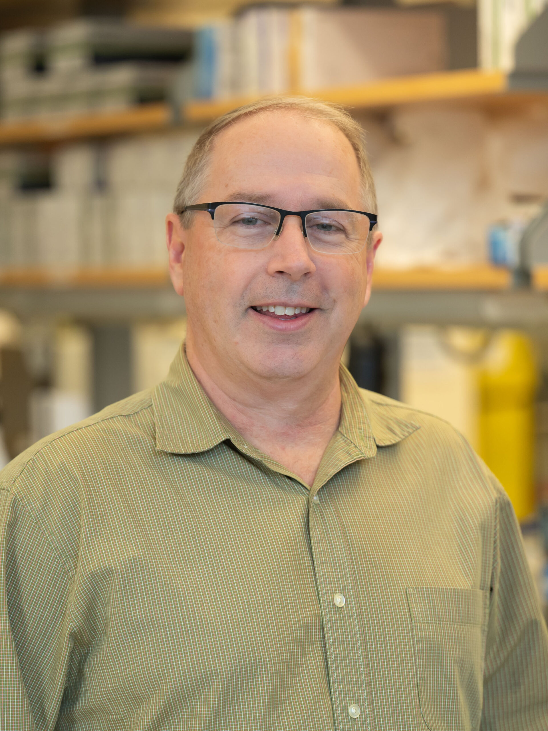 Headshot of Jeff Holt. He is standing in a lab, wearing a green button down shirt and glasses