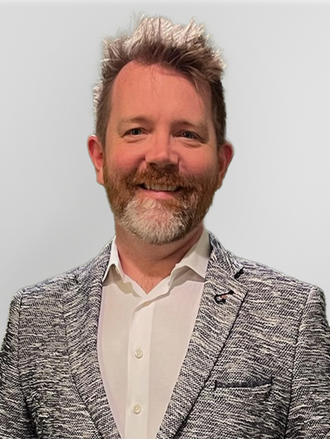 A professional headshot of a man, Michael Conn. He is wearing a white button down and gray jacket and he has short gray hair and a beard. He is smiling and looking straight ahead.