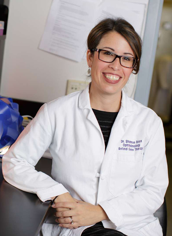 A professional headshot of a woman, Shannon Boye, wearing a white lab coat and learning her arm on a table. She has a wide smile, brown hair, and wears dark rimmed glasses.