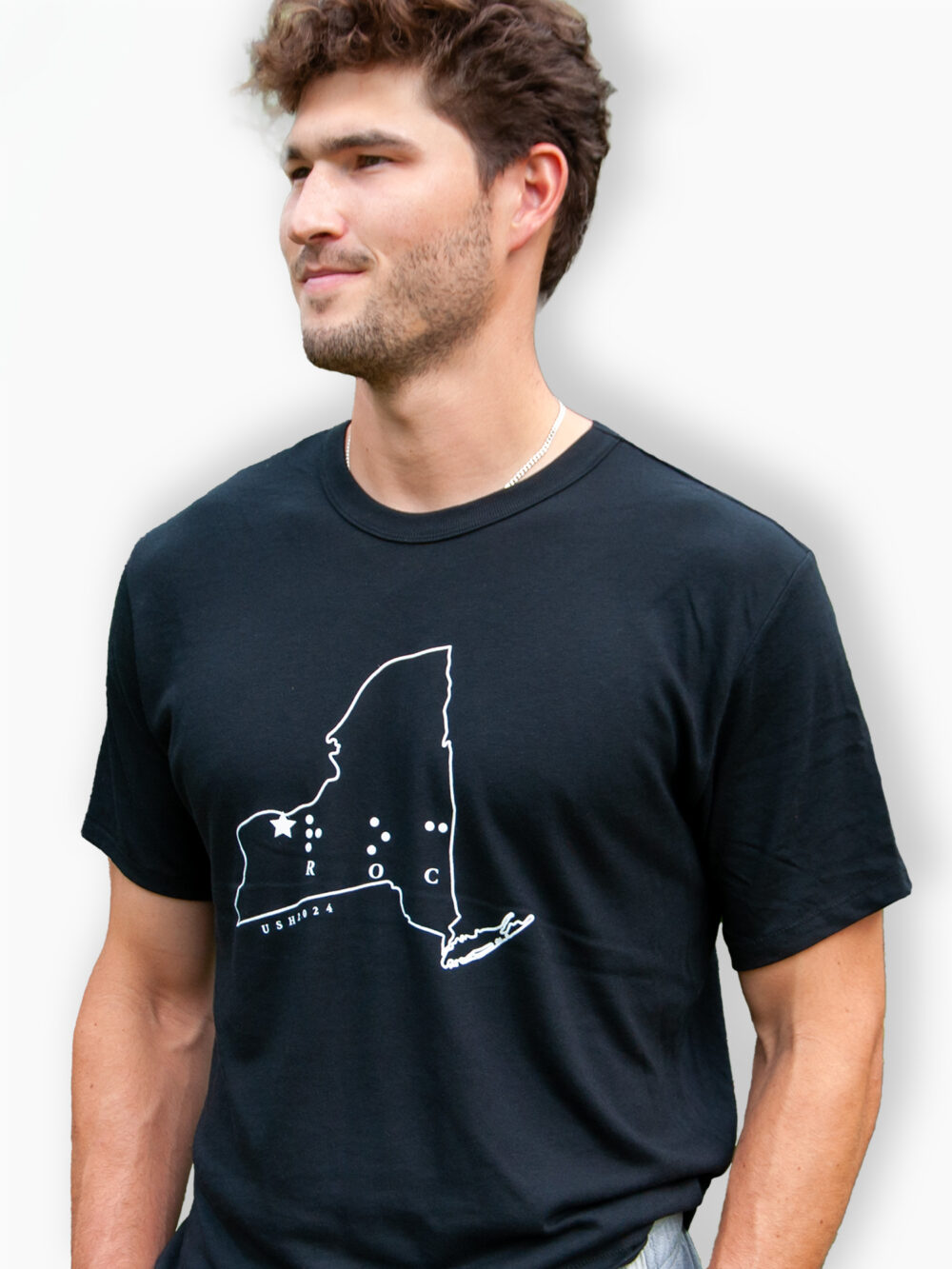 A young man is wearing a black t-shirt with a white line drawing of New York state with the letters "ROC" in braille inside the state outline and USH2024 written on the edge of the outline