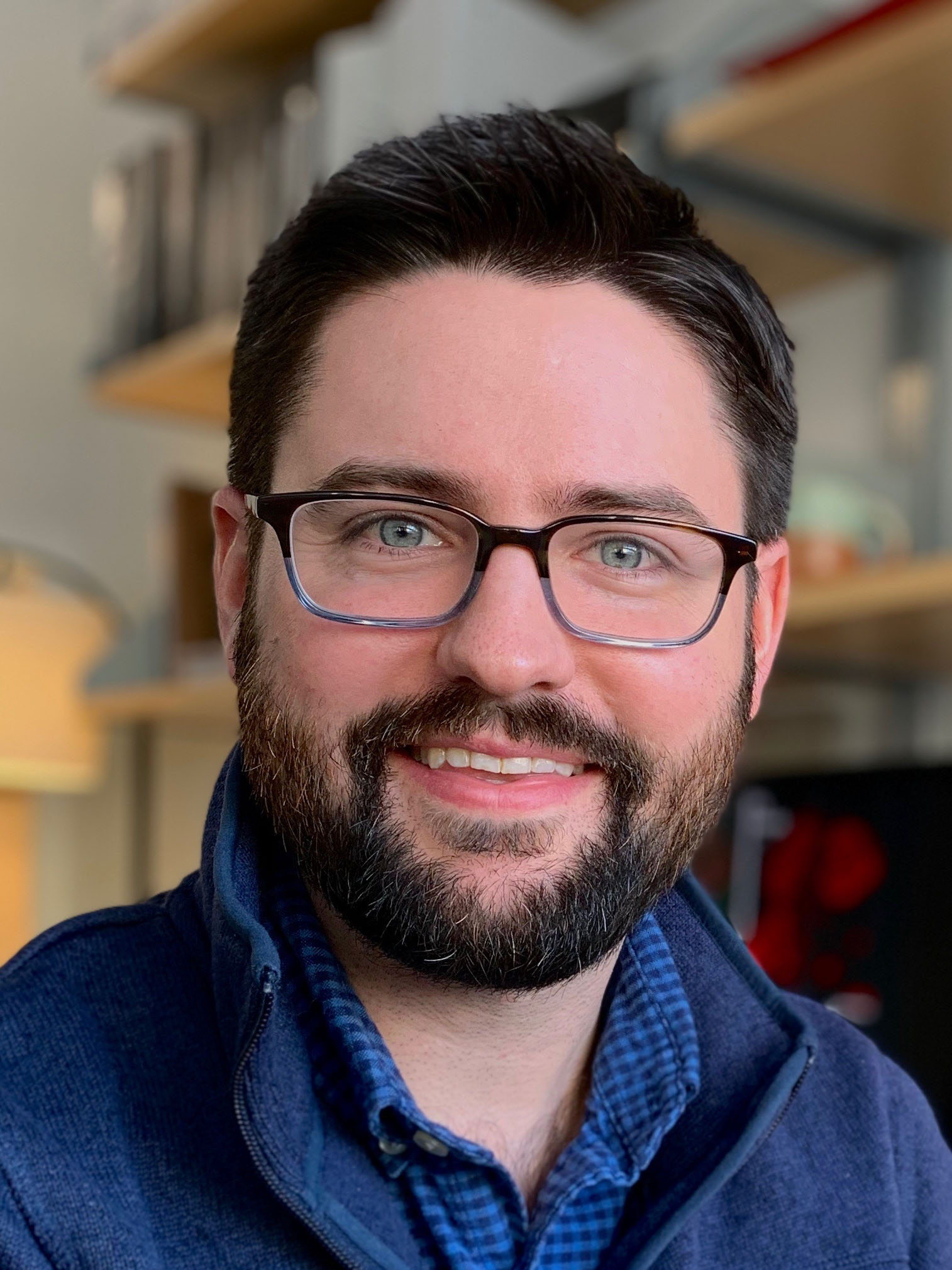 Headshot of Karl Koehler. He has dark hair. a dark beard, and wears glasses. He has on a blue collared shirt and he is smiling.