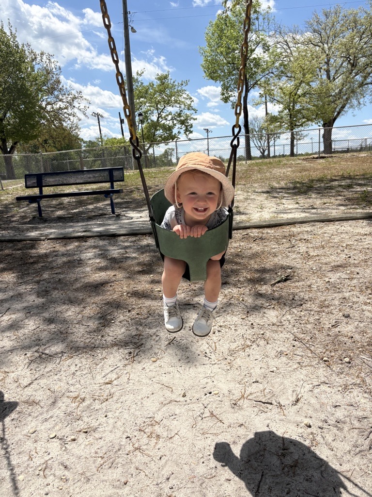 Joey is outside wearing a big hat and swinging on a swingset