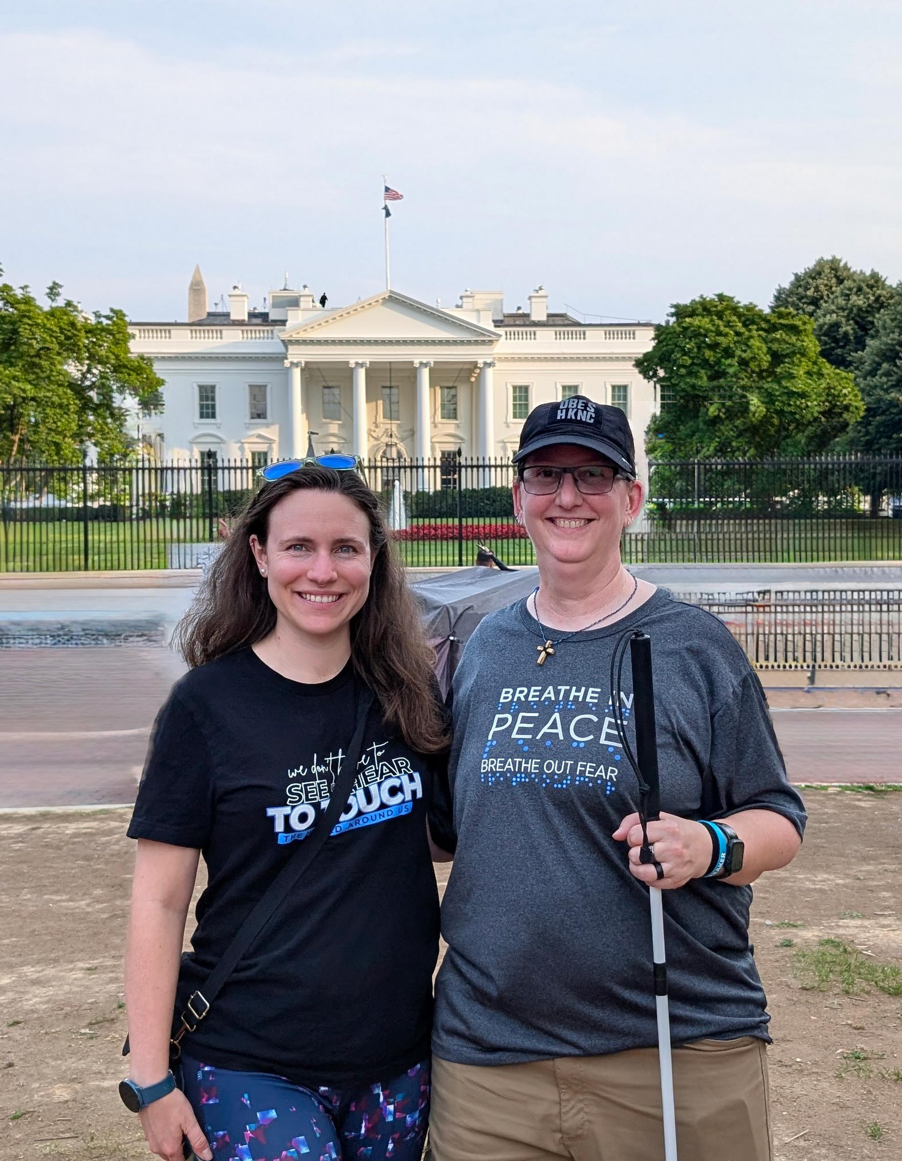 Picture 4-2 Laurie (right) is wearing a dark baseball cap, glasses, and a dark gray t-shirt that says, “Breathe in PEACE, Breathe out fear.” She is holding a white cane in her right hand and standing beside McKenna, Laurie's SSP, who has long brown hair and is wearing a black t-shirt that reads, “We don’t see barriers. TO TOUCH is to turn things around,” and colorful leggings. The Washington Monument is visible in the background behind the White House, along with green trees and a blue sky.
