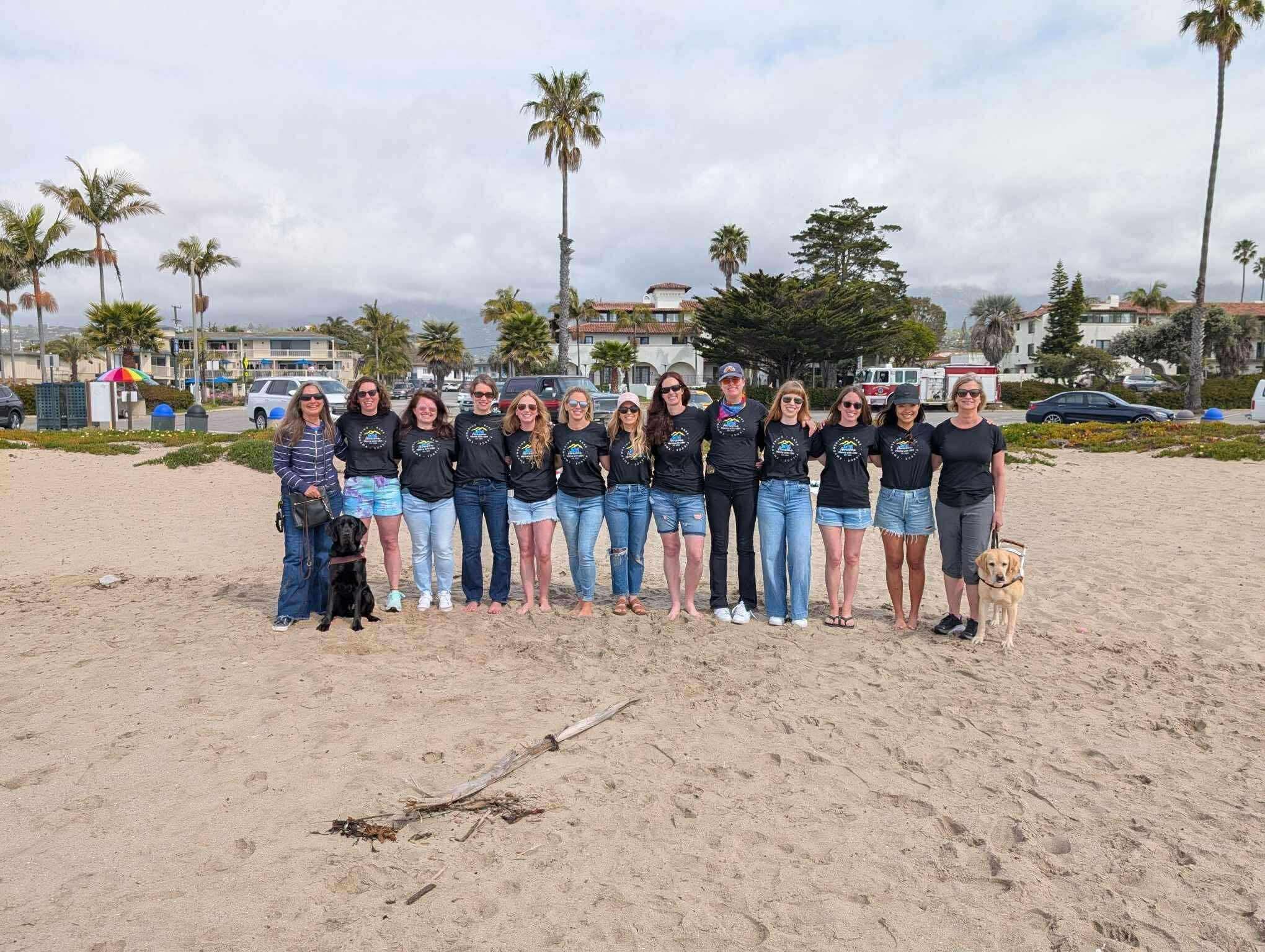 USH Community Meagan Moore A group of thirteen women stands together on a sandy beach during the 2025 Awesome Moms with Ush Retreat in Santa Barbara. Most wear matching black retreat shirts and denim, smiling with arms around each other. A black guide dog and a yellow guide dog sit at each end of the group. Palm trees and coastal buildings line the background under a cloudy sky, capturing a moment of connection and community.