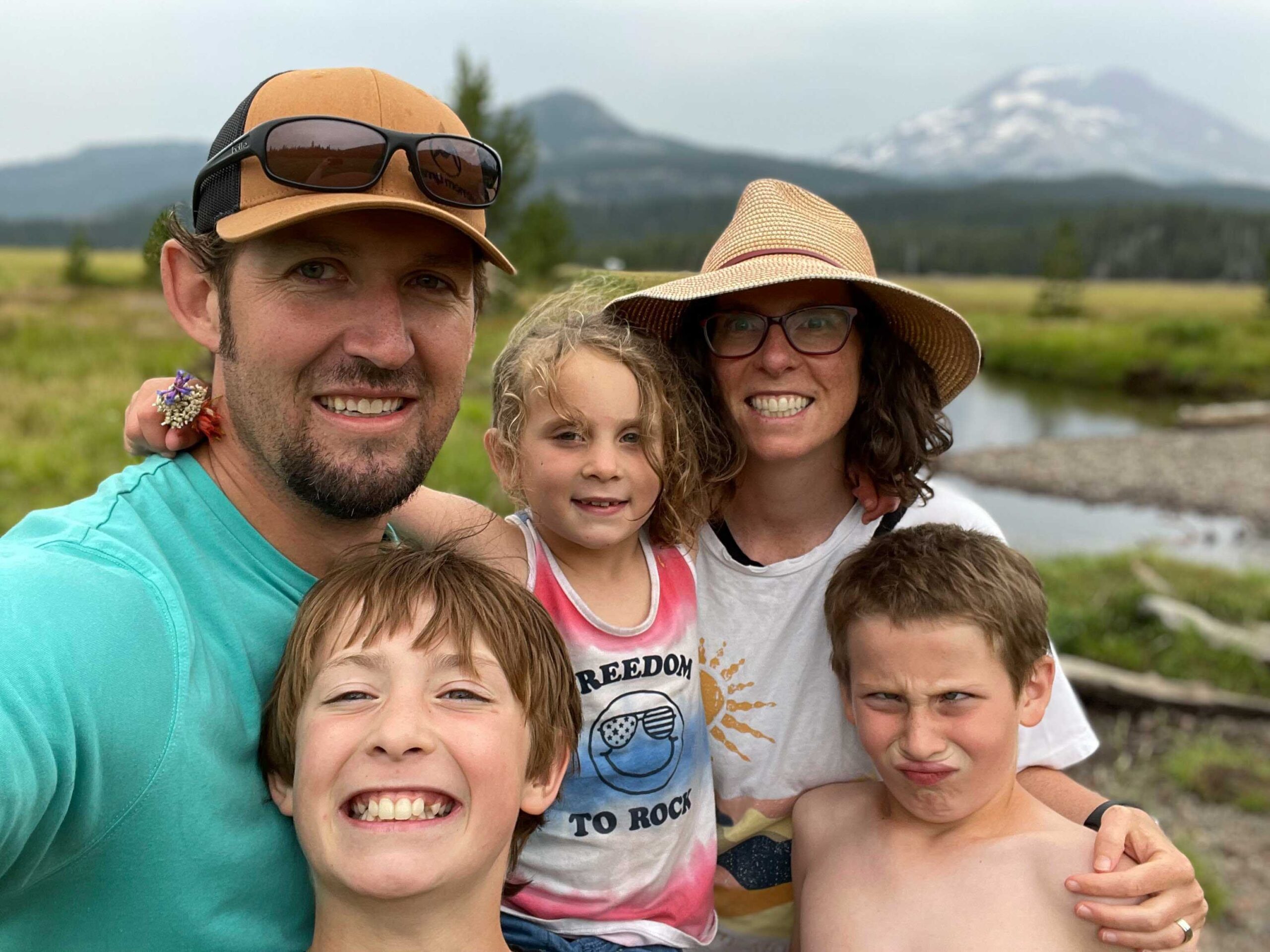 Meagan Moore family Photo Meagan and Joel stand with their three kids in a grassy field with a stream and snow-dusted mountains in the background—a place they frequent and enjoy often. Meagan wears a straw hat and glasses, and Joel wears a brown cap and teal shirt. Their daughter, in a “Freedom to Rock” tank top, holds a small bouquet of wildflowers and smiles. In front, their two shirtless sons pose—one grinning, the other making a playful face. The overcast sky and scenic mountain setting frame this joyful family moment.