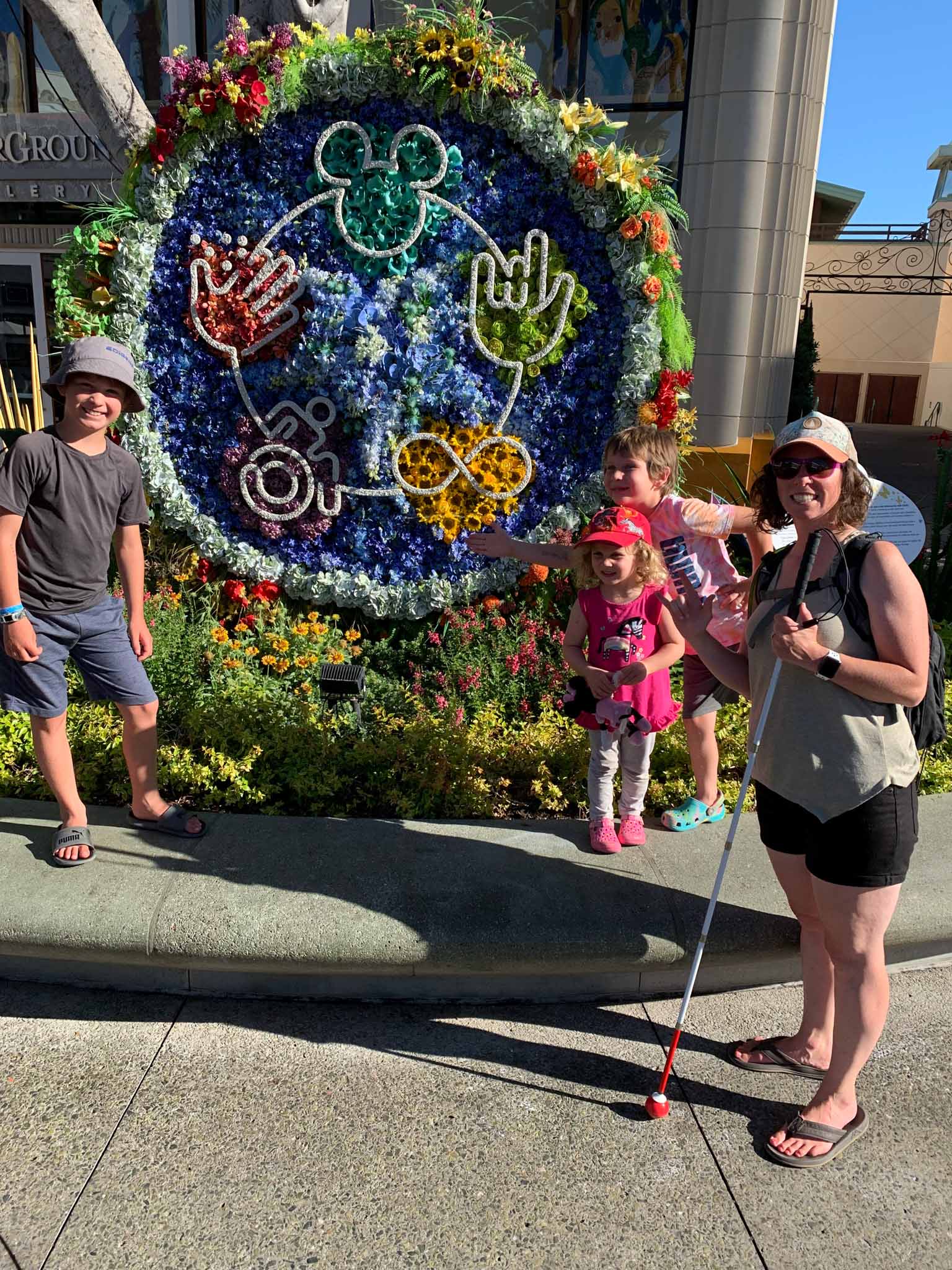 Meagan and Kids Meagan stands with her three kids in front of a large, colorful flower display at Disneyland. The circular floral arrangement features symbols of inclusion made from flowers, including a wheelchair, an infinity sign, ASL hand signs, and Mickey ears. Meagan, holding a white cane with a red tip in one hand, makes the “I love you” sign in American Sign Language with the other. She smiles brightly, wearing sunglasses and a hat. Her three kids, dressed casually and also smiling, pose beside her in the sunny, flower-filled setting.