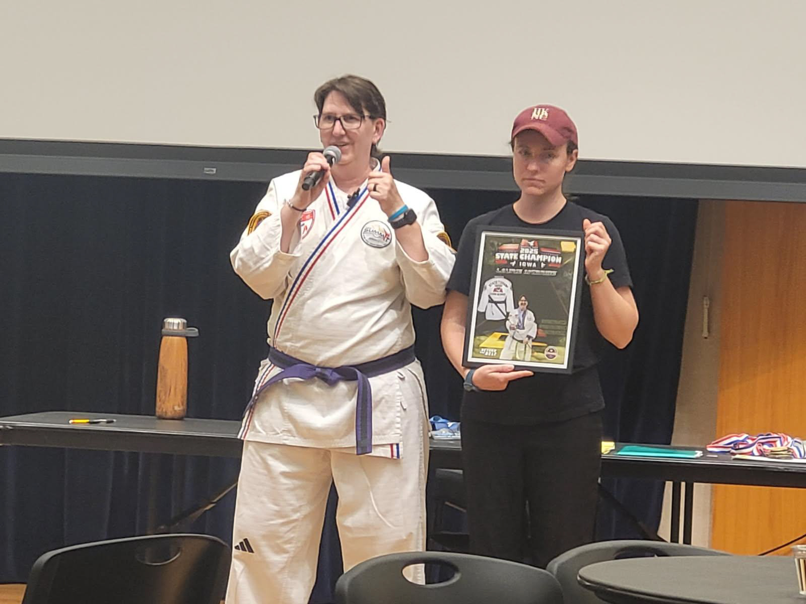 Laurie McBride Legacy in Action Tournament Laurie is wearing her Taekwondo Gi (uniform) with a purple belt. She holds a microphone and is mid-speech, using expressive hand gestures and smiling proudly. On her left is McKenna, her Support Service Provider (SSP)