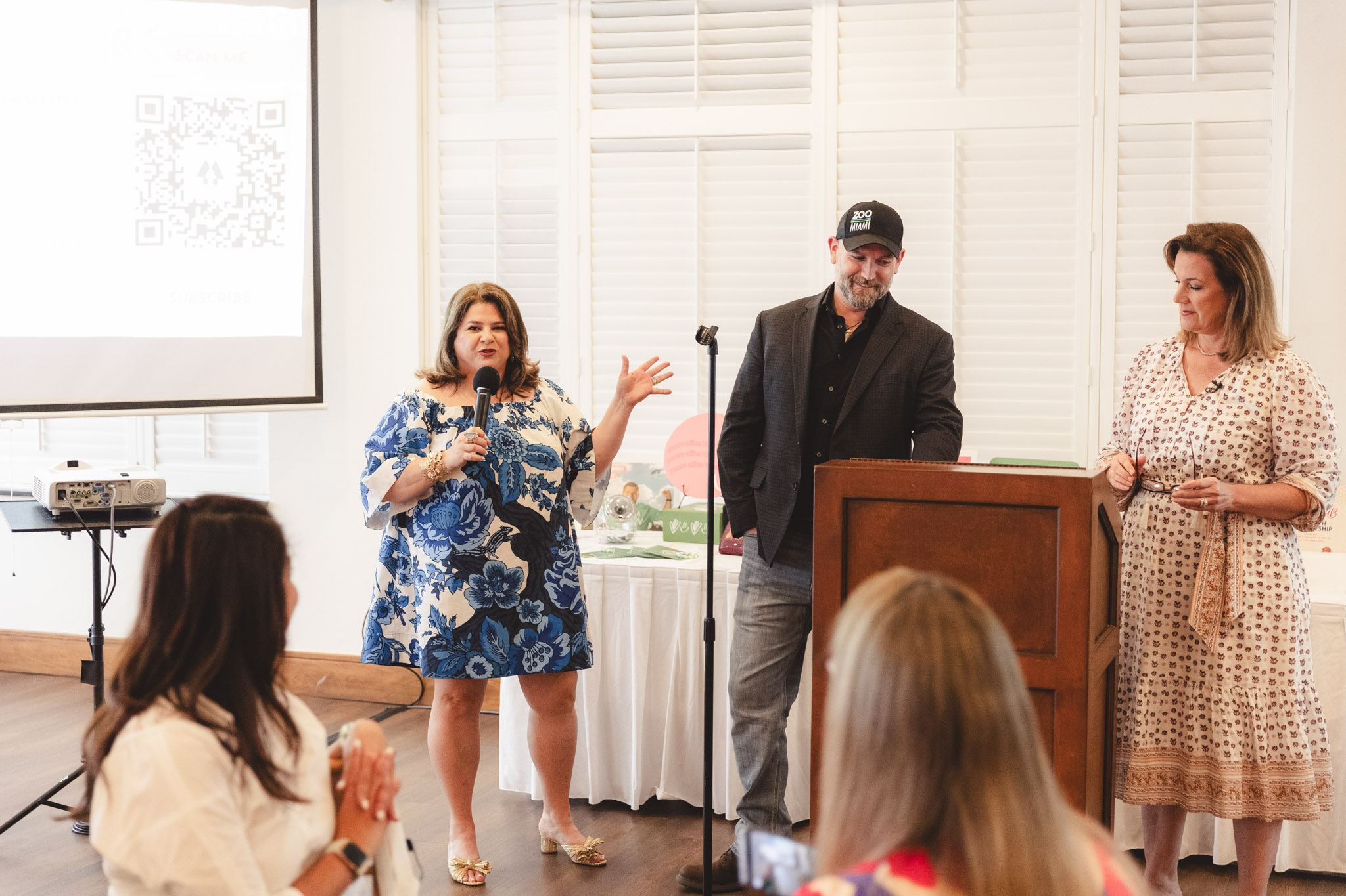 Samantha speaks in a mic during an event for 305 Hive Media. She wears a blue floral dress and stands at the front of the room near a podium.