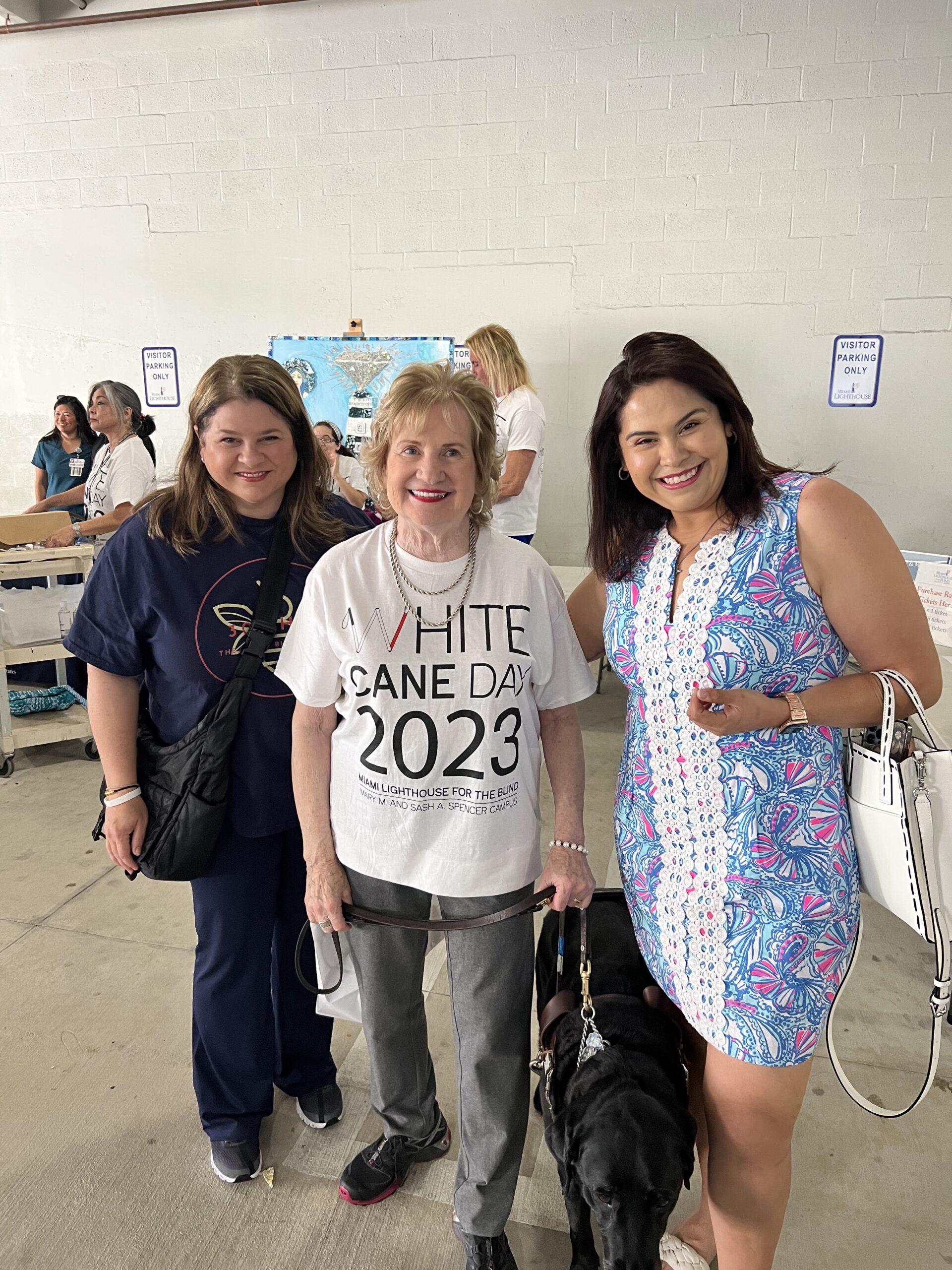 Samantha stands with two other women during a white cane day. They all look at the camera smiling and one woman wears a white t-shirt that says "white cane day 2023"