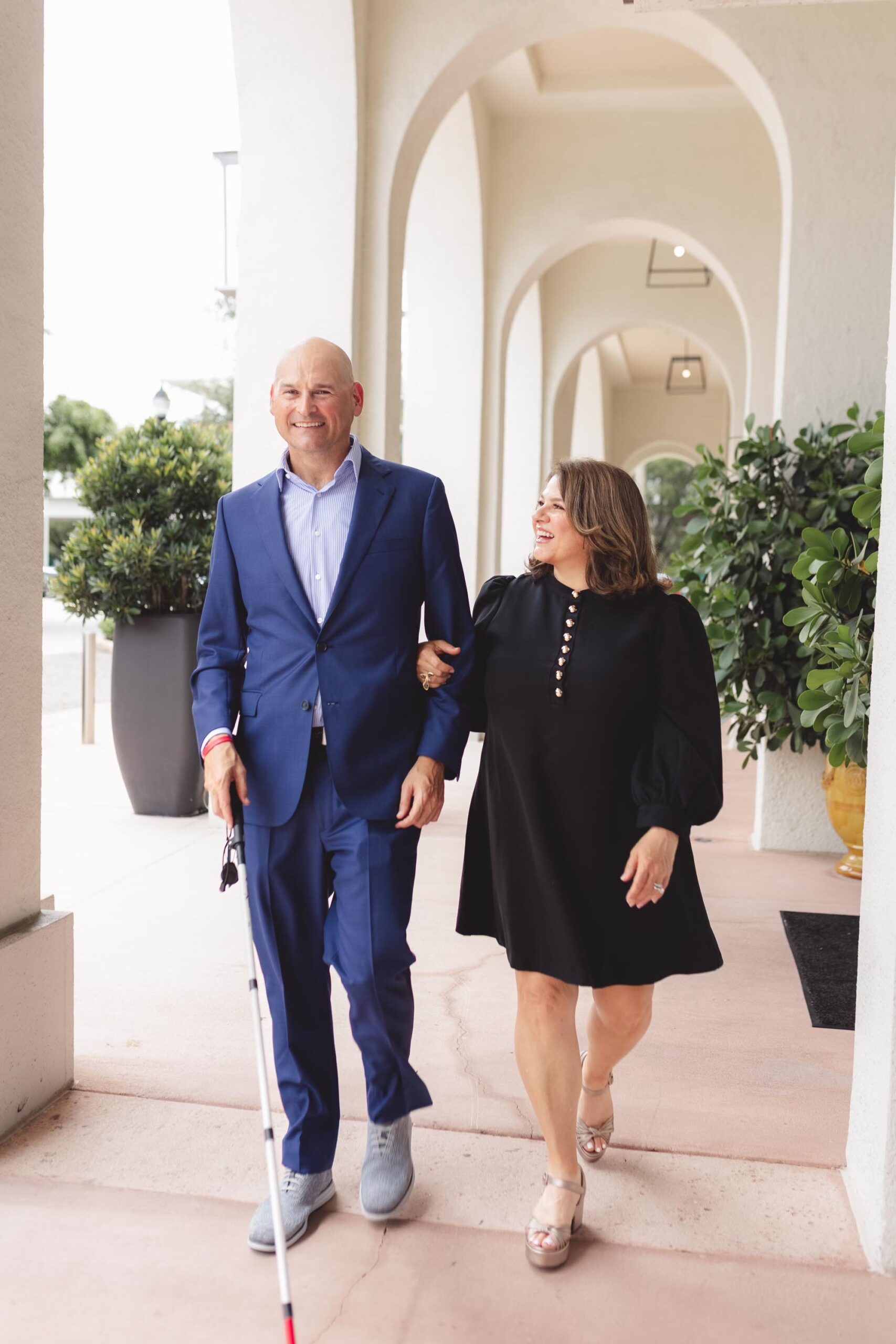 Samantha walks arm and arm with her brother Michael. Her brother wears a blue suit and is using a cane and Samantha wears a long sleeve black dress.