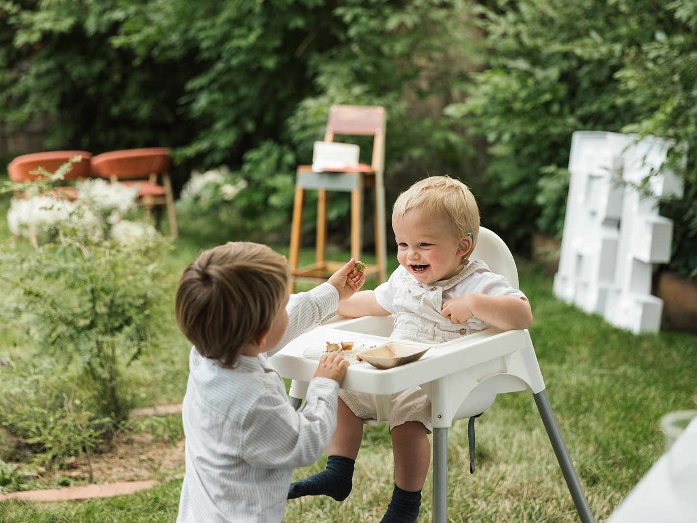 Olivier and Brother Olivier sits in a high chair as his brother hands him something. They are outside in a beautiful green landscape.