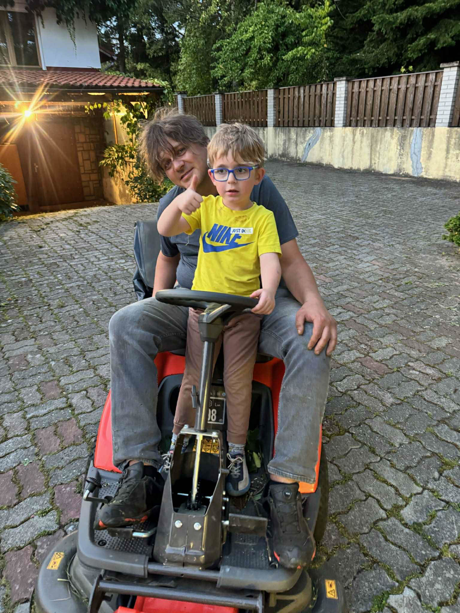 Stepanek sits in the drivers seat of a lawnmower giving a thumbs up. A man sits behind him. 
