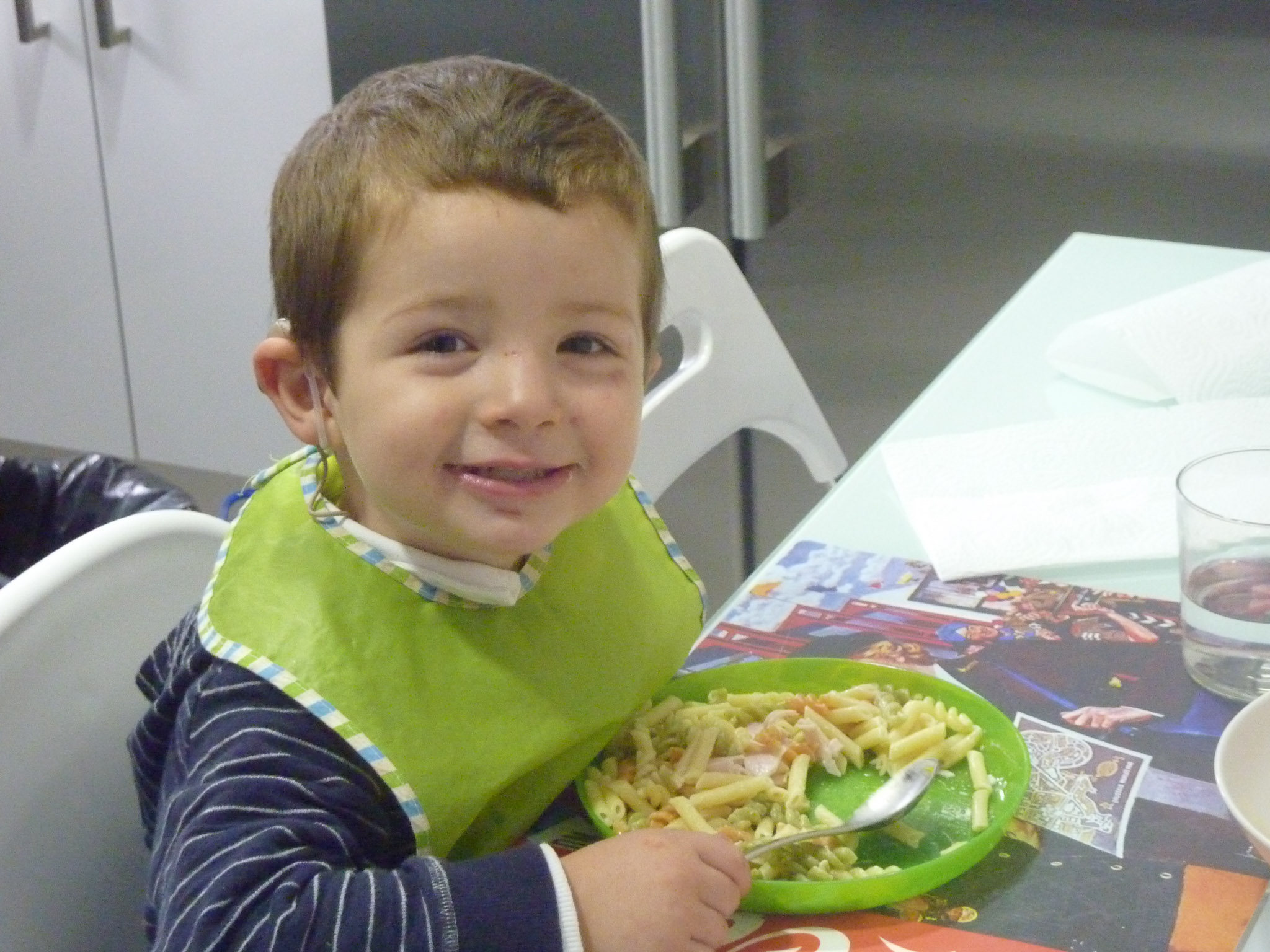 A photo of Daniel sitting at the table wearing a green bib and eating. He looks at the camera and smiles