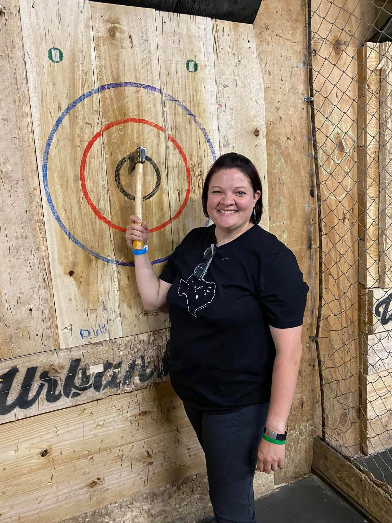 A smiling adult woman stands indoors in front of a wooden target used for axe throwing. The target is made of light-colored wood with painted rings: a large blue circle, a red circle inside it, and a small black bullseye at the center. An axe is embedded in the bullseye. The woman is standing to the right of the target, facing the camera. She has short, dark hair and is wearing a black T-shirt and dark pants. She holds the axe handle upright with one hand, her arm bent at the elbow, showing that the axe has landed in the center of the target. A pair of glasses hangs from the collar of her shirt. The setting appears to be an axe-throwing venue, with rough wooden walls and a chain-link safety fence visible to the right. The overall mood is cheerful and proud, as if she has just made a successful throw.