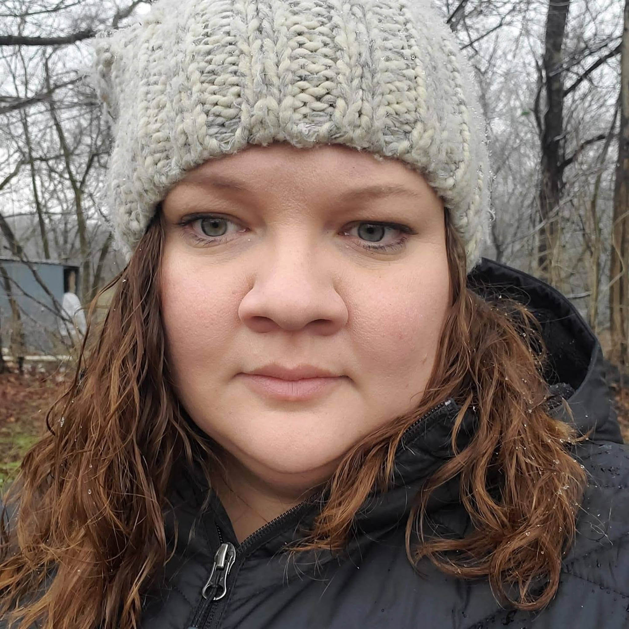 A close-up outdoor portrait of an adult woman facing the camera. She is wearing a light gray, thickly knitted winter hat pulled down over her forehead and a black, zip-up jacket suitable for cold weather. Her hair is medium-length, brown, and slightly wavy, falling around her face from beneath the hat. Her expression is calm and neutral, with relaxed eyes and closed lips. The background shows a quiet, wintry setting with bare tree branches and muted colors, suggesting late fall or winter. Small droplets or light moisture are visible on her hat and jacket, giving the impression of mist, drizzle, or melting snow. The overall feeling of the image is quiet, cool, and still.