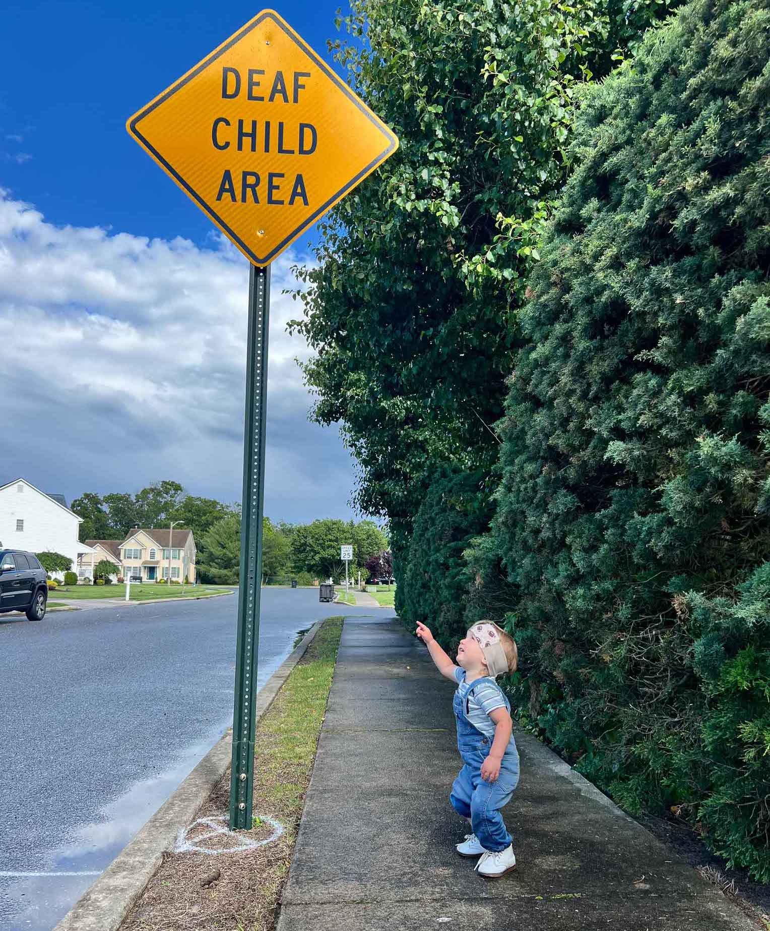 A photo of a young boy standing on the sidewalk. He is looking up and pointing to a yellow sign that says Deaf Child Area