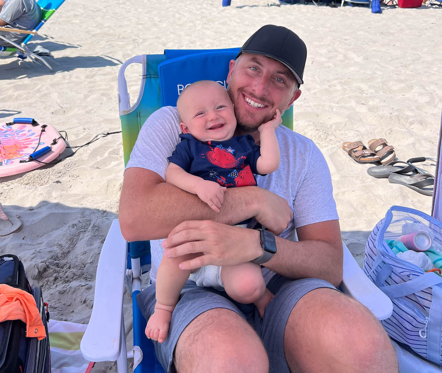 Michael Poalise sits in a chair on the beach holding Joey. They are both smiling wide.