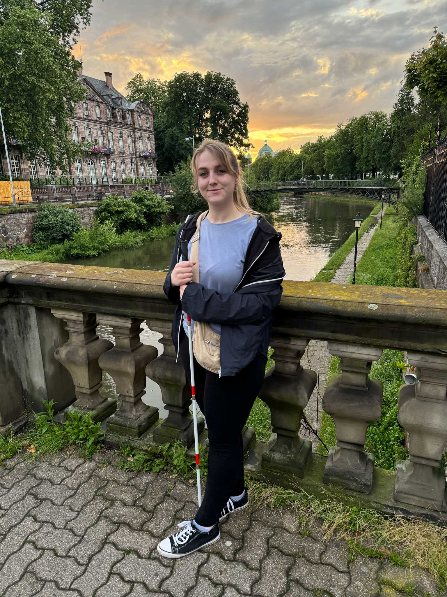 Chloe poses against a railing on a bridge with a beautiful sunset behind her and greenery.