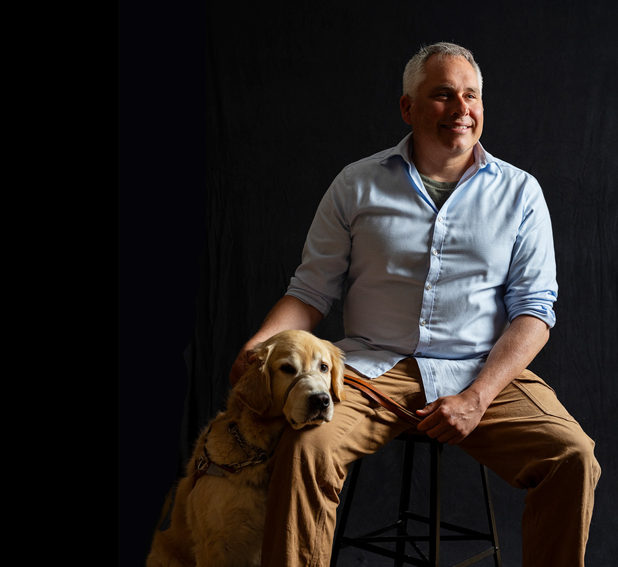 A portrait of a man seated with his dog beside him, resting his head on the man's leg. The man wears a button down shirt and looks off to the side. He is half illuminated by light and half in shadow.