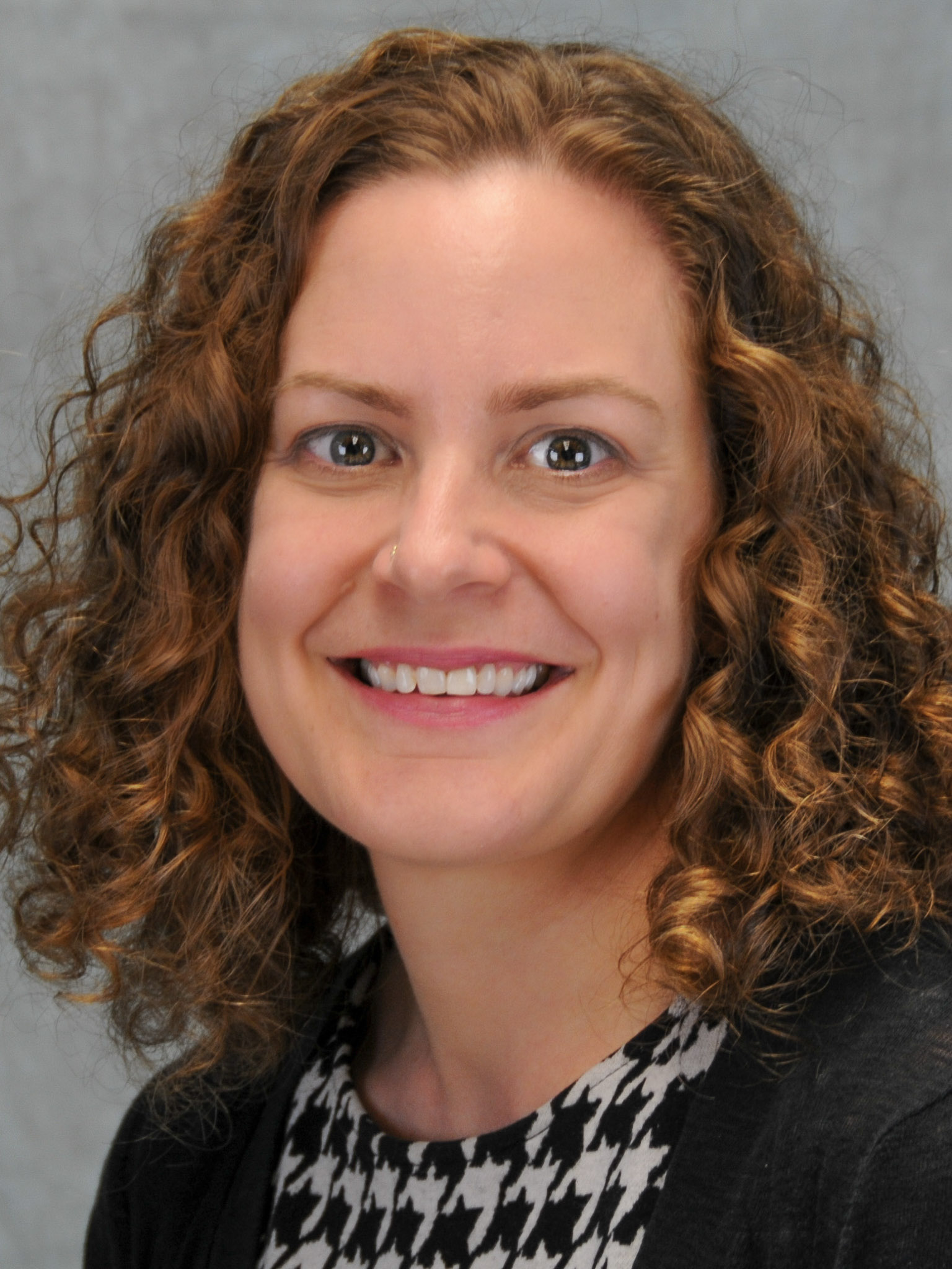 A close up headshot of a woman with curly red hair is smiling and looking straight ahead. She wearing a black and white checkered top and there is a gray background behind her.