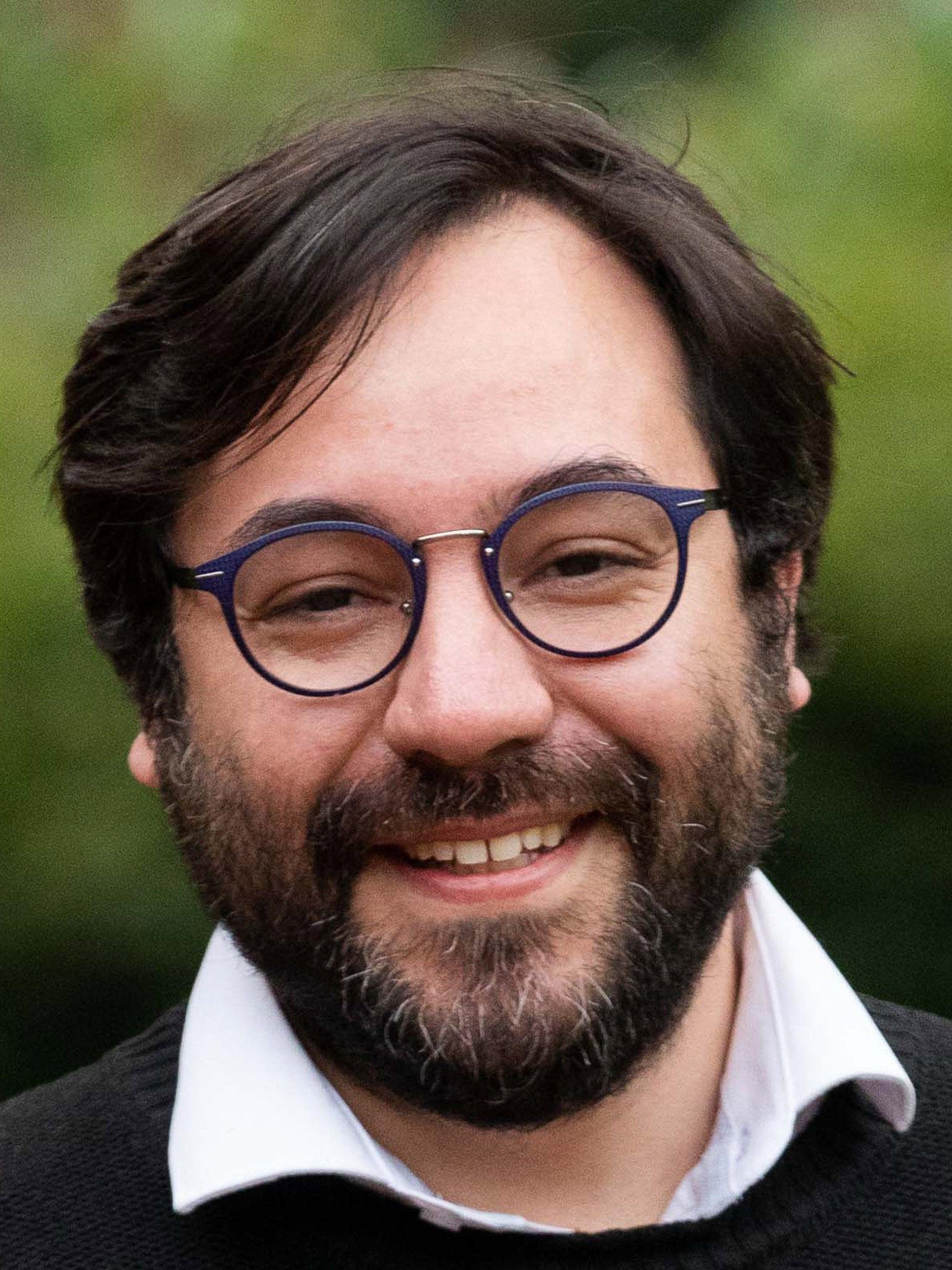 A close up image of a man with dark hair, a dark beard, and round glasses. The man smiles and looks straight ahead. He wears a white colored shirt and a black sweater.