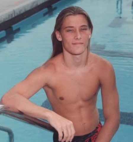A young, long-haired Nathan Stockton is pictured poolside during his high school freshman swim team picture day in 1995, highlighting a lifelong passion for swimming that will culminate in his participation in the Usher Syndrome Society’s Ush X Swim Alcatraz event in October 2026