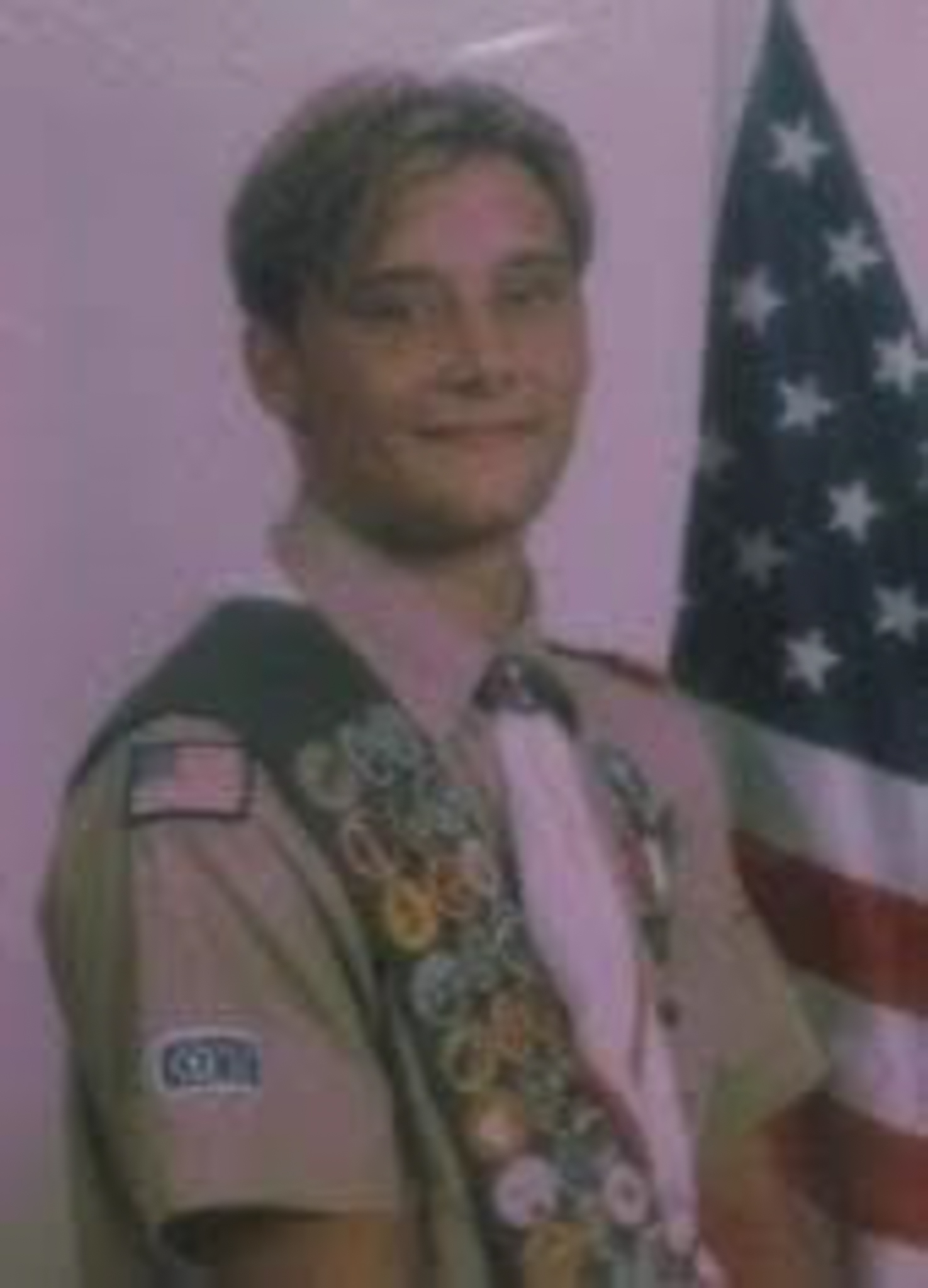 An 18-year-old Nathan Stockton poses proudly next to an American flag during his 1998 Eagle Scout ceremony, wearing his full uniform and merit badge sash, which includes the Wilderness Survival badge he uniquely earned by sleeping alone in the wild despite a nearby bear sighting