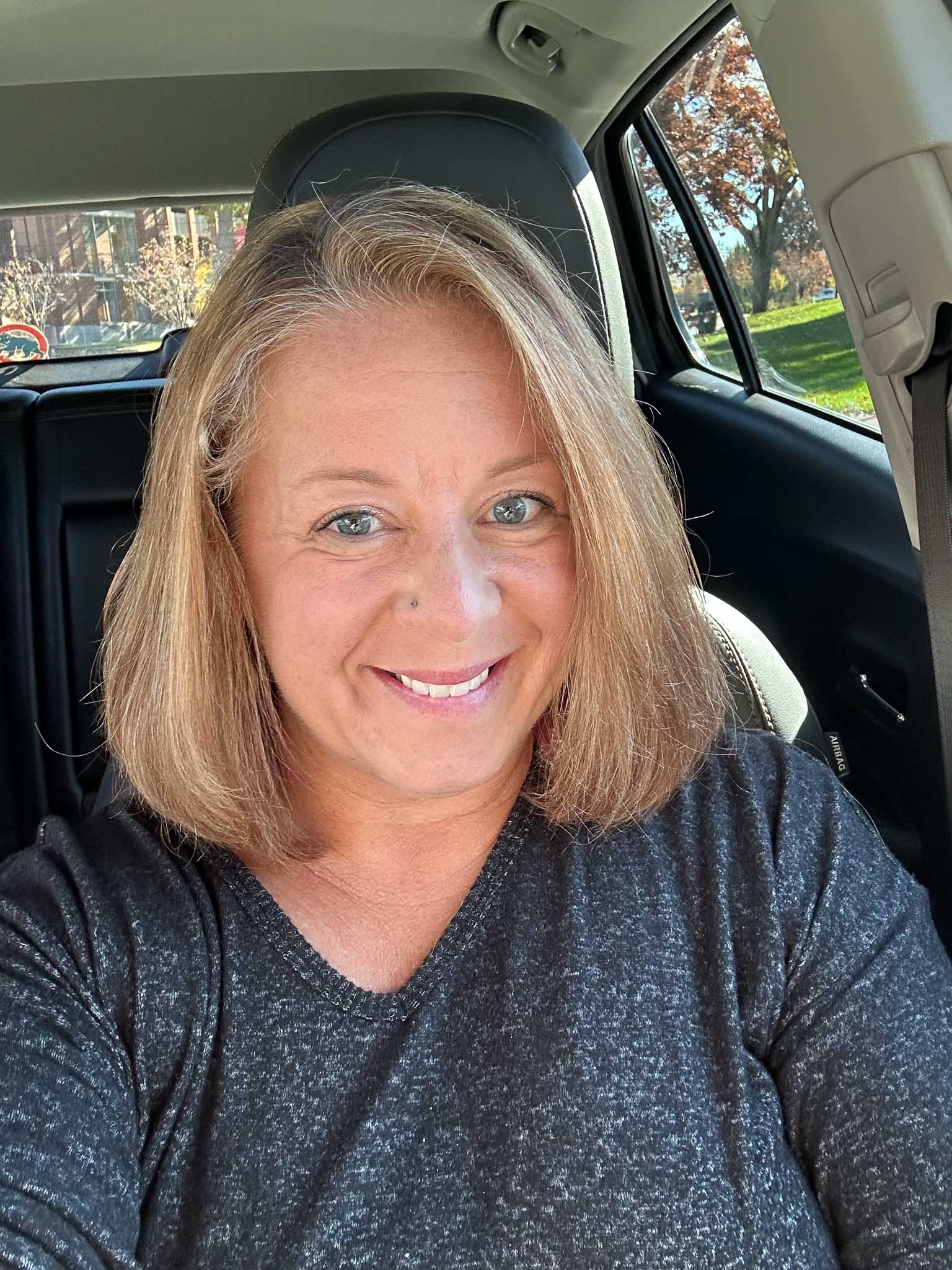 A selfie photo of a woman with shoulder length blonde hair sitting in a car. She is smiling and looking at the camera and wears a gray t-shirt.
