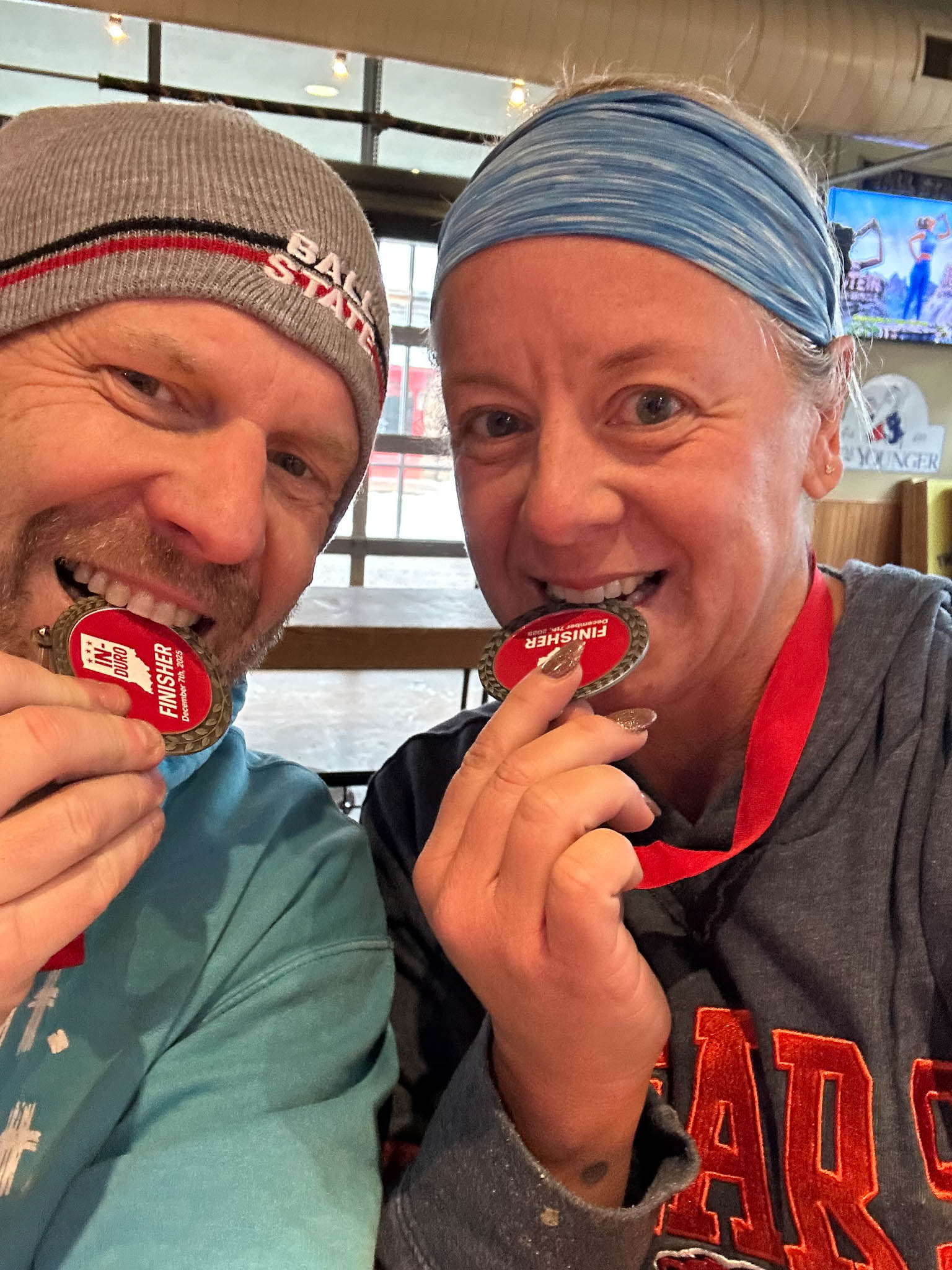A close up image of a man and a woman taking a selfie. They are both wearing beanie hats and biting a medal with a smile on their faces.