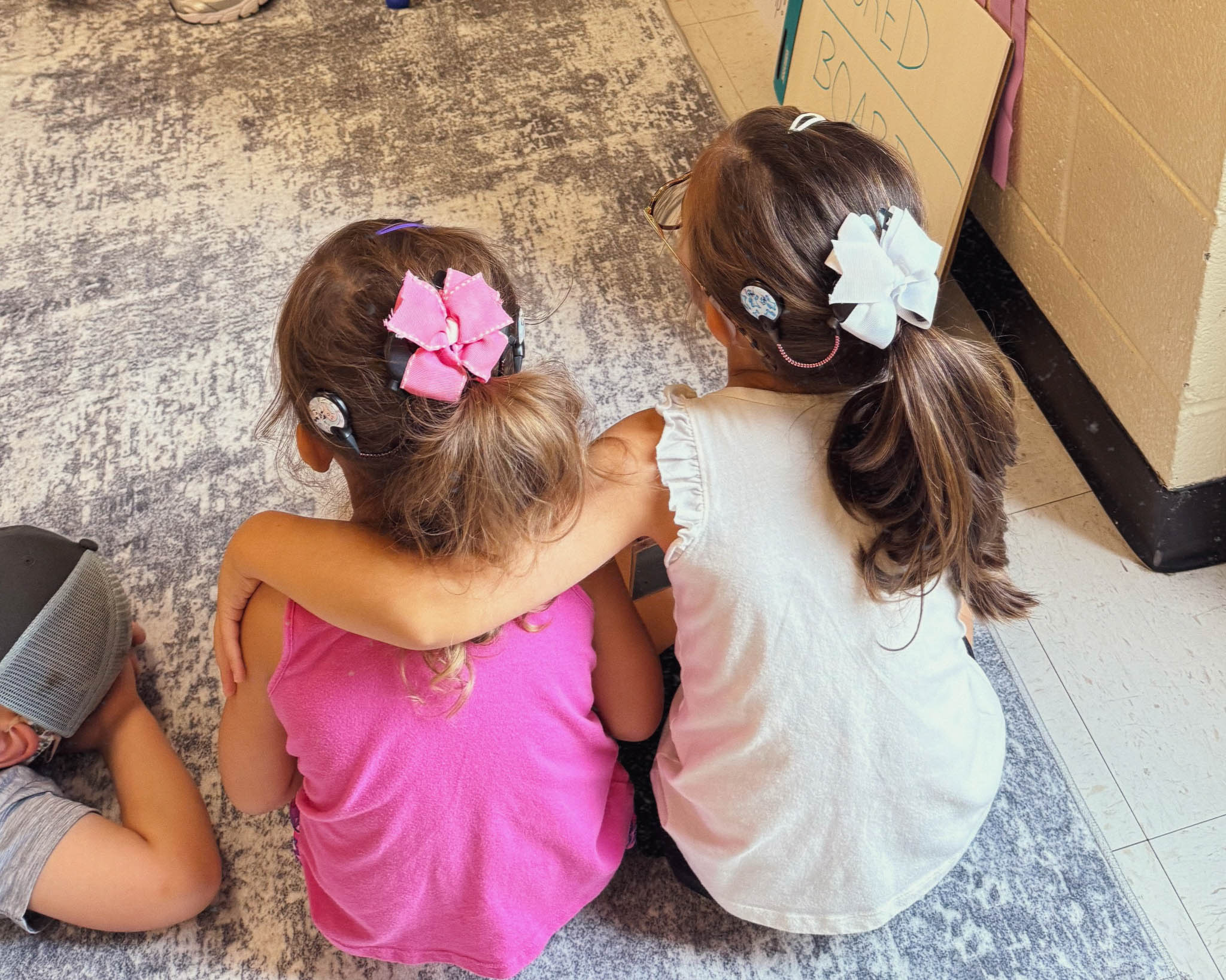 A photo of two young girls sitting on the ground side by side with their back to the camera. One of the girls has their arm around the other's shoulders.
