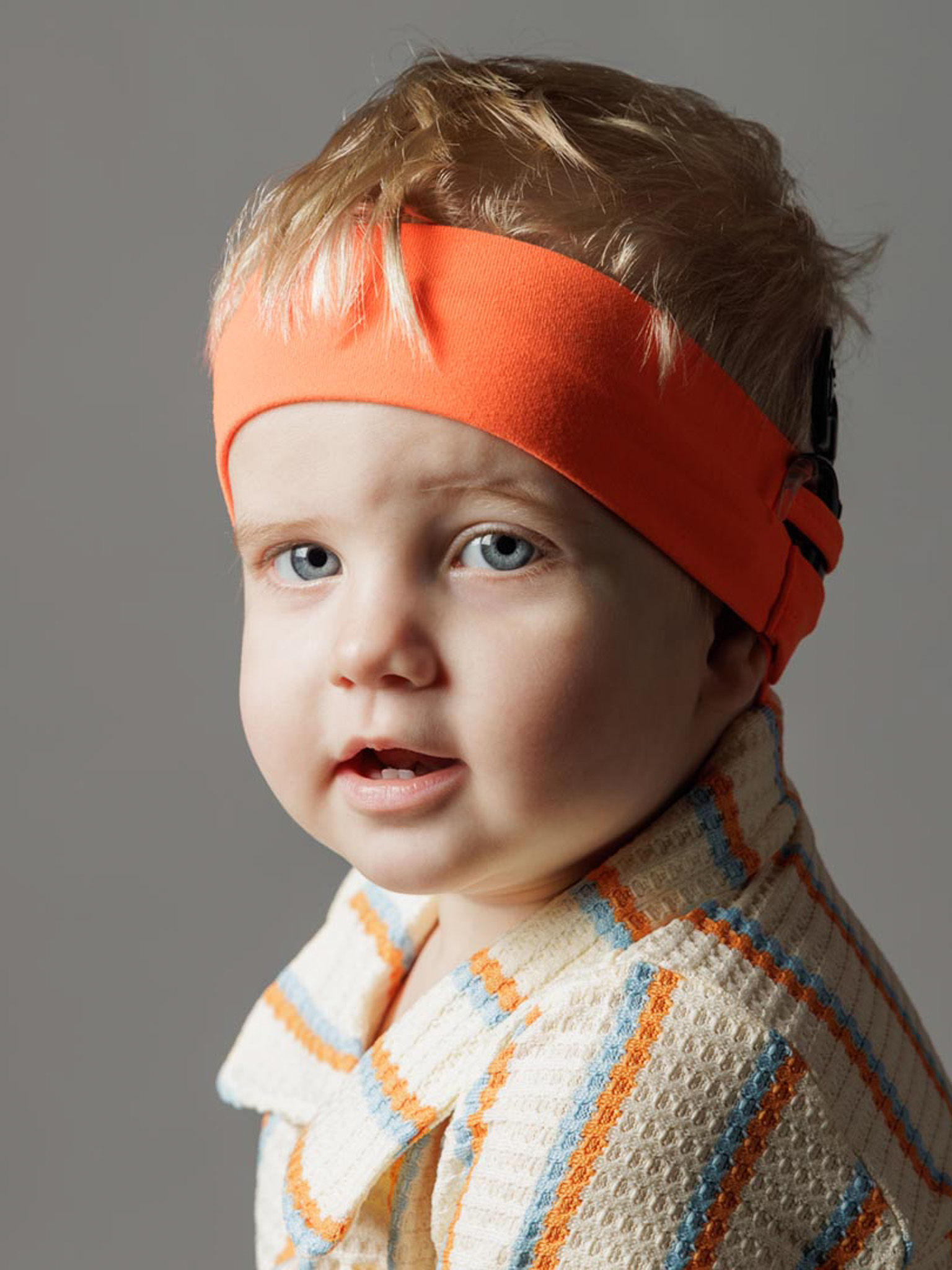 A young boy with light blonde hair wearing a thick orange headband. He looks directly at the camera. 