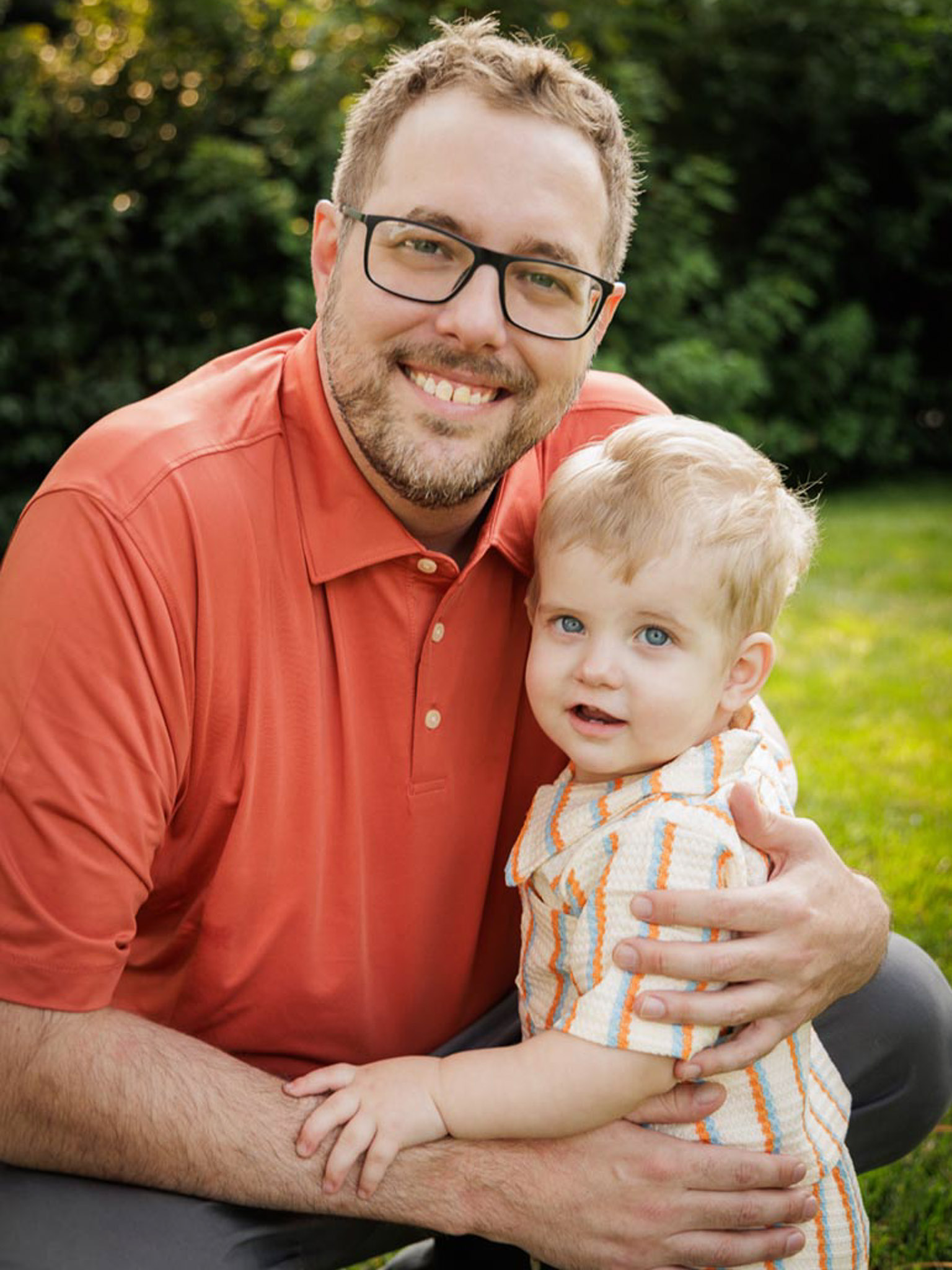 A young boy is held by an older man. They are both looking at the camera smiling and holding one another. The man wears an orange polo shirt and the boy wears a striped shirt.