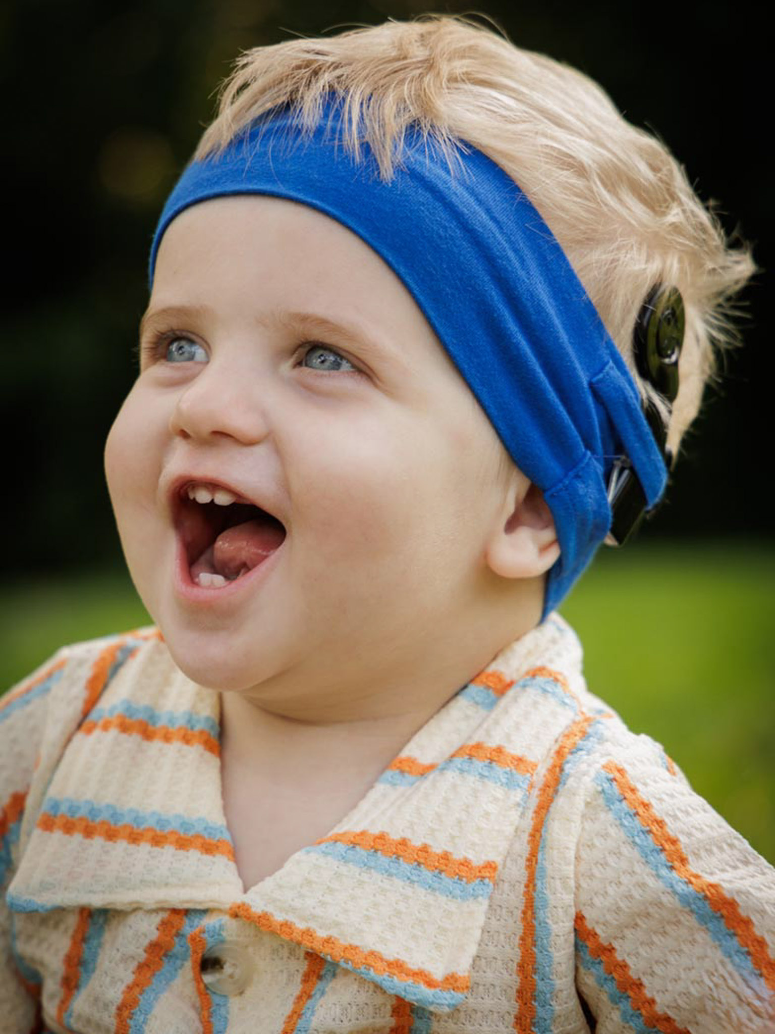 A young boy with light blonde hair wearing a blue headband is outside and smiling wide