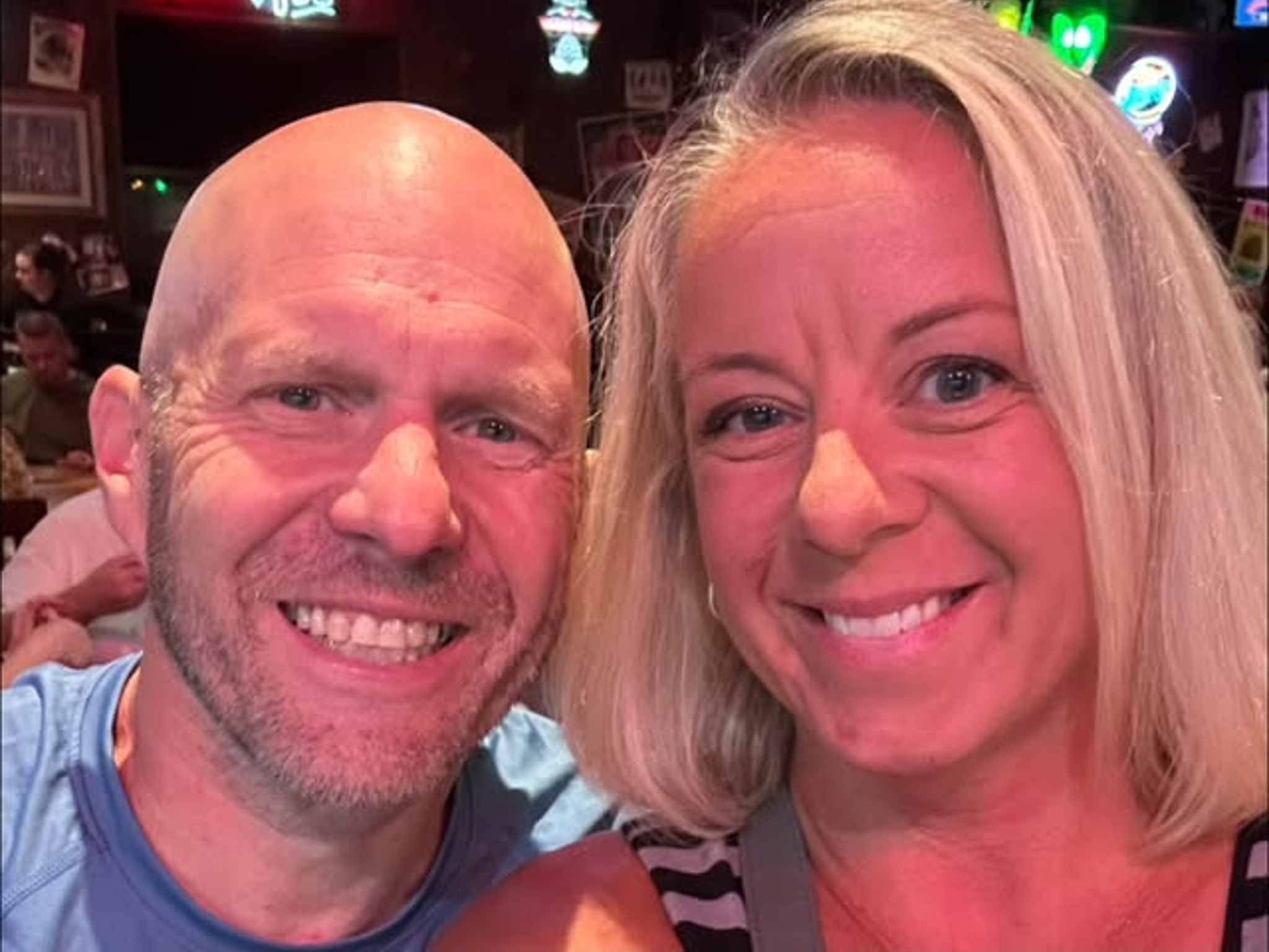 A close up image of a man and woman who are both smiling wide. The woman has short blonde hair and the man has no hair.