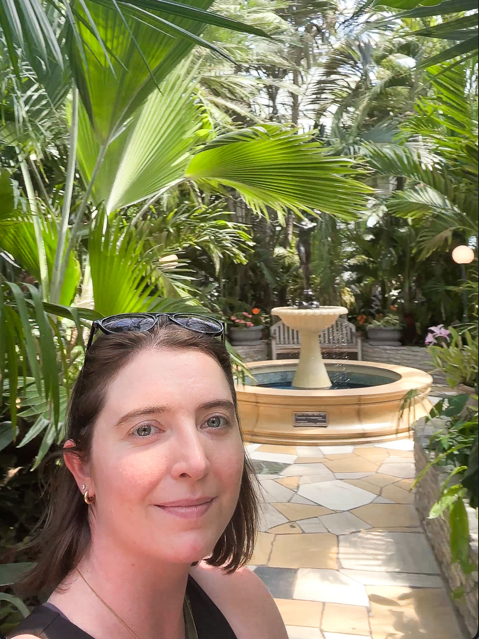 A young woman with short brown hair standing in a room with a fountain and lots of plants with large leaves behind her.