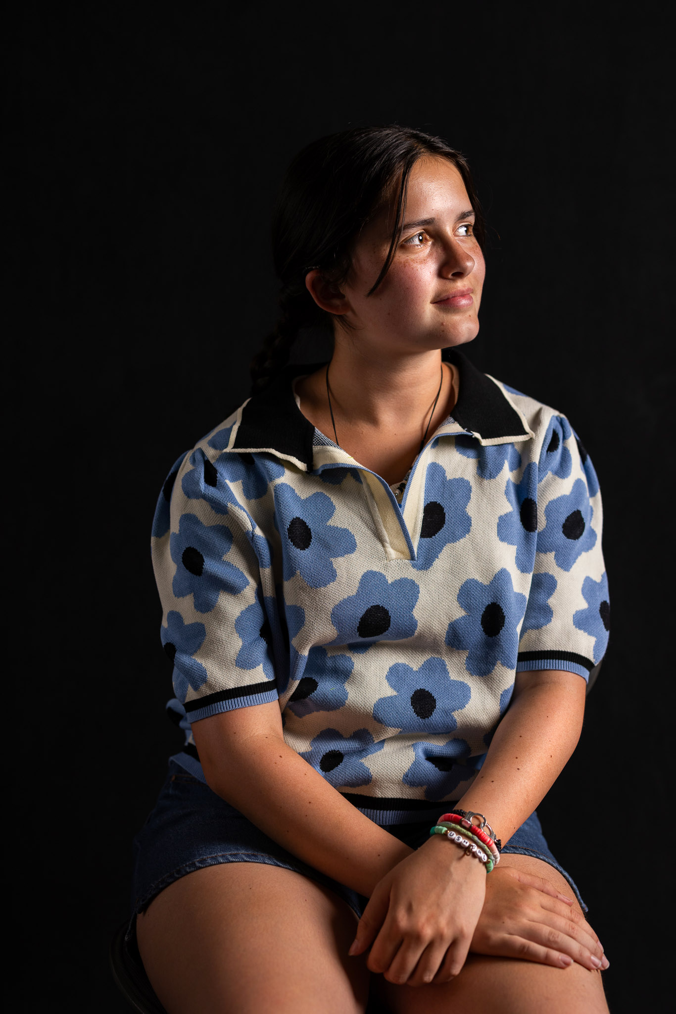 A young girl with brown hair pulled back in a ponytail is seated, looking off to the side with her arms crossed on her lap. She is illuminated by light, with a dark black background behind her.