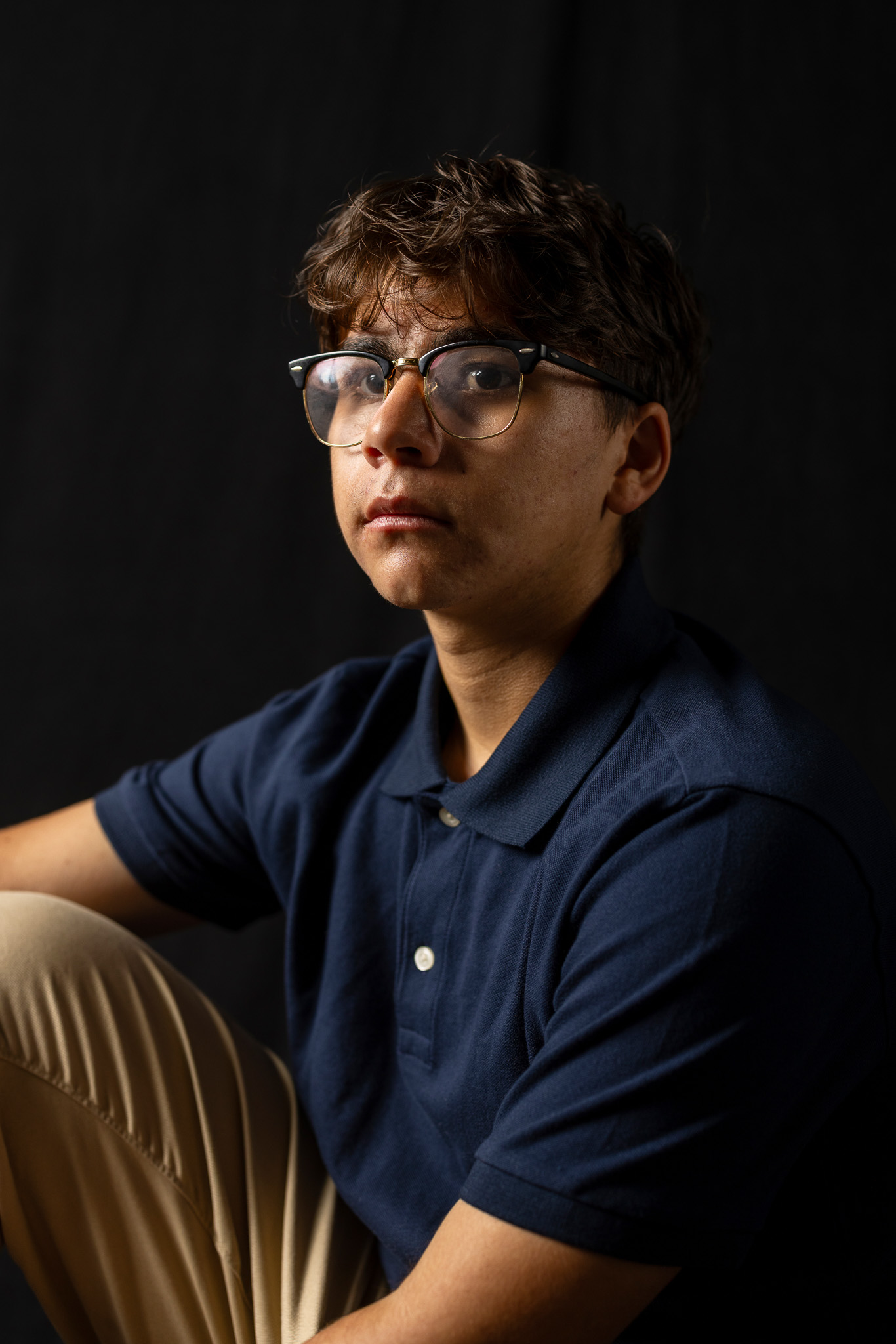 A photo of a young boy with brown curly hair and glasses looking off to the side. He is half illuminated by light and half in shadow.
