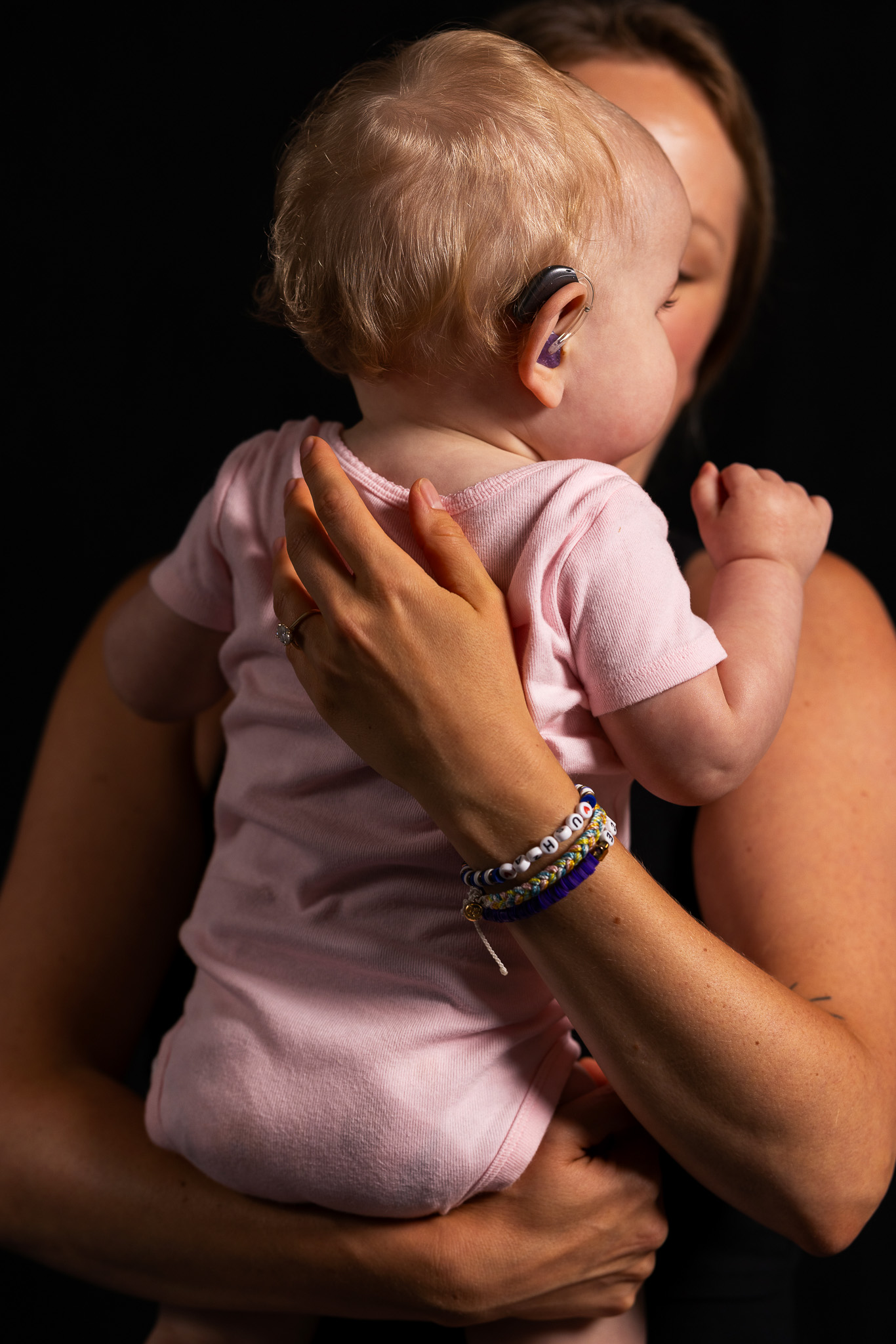 A photo of a young baby being held in their mothers arms. The baby wears hearing aids and is illuminated by light, with a dark black background behind them.