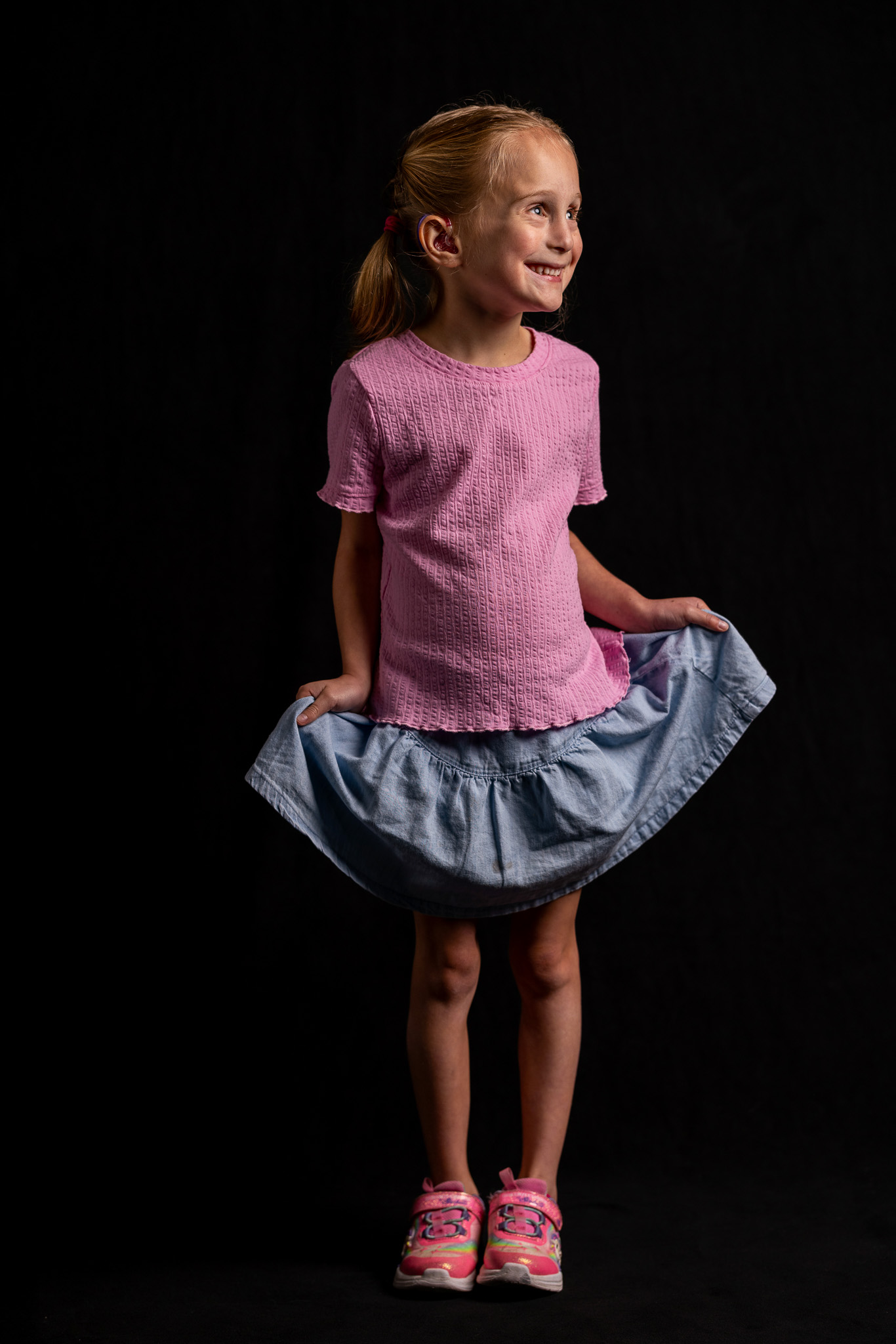 A young girl stands in front of a black background. She is smiling and looking off to the side, while holding her miniskirt out to the side with both hands. She is illuminated by light, with half of her face in shadow. 