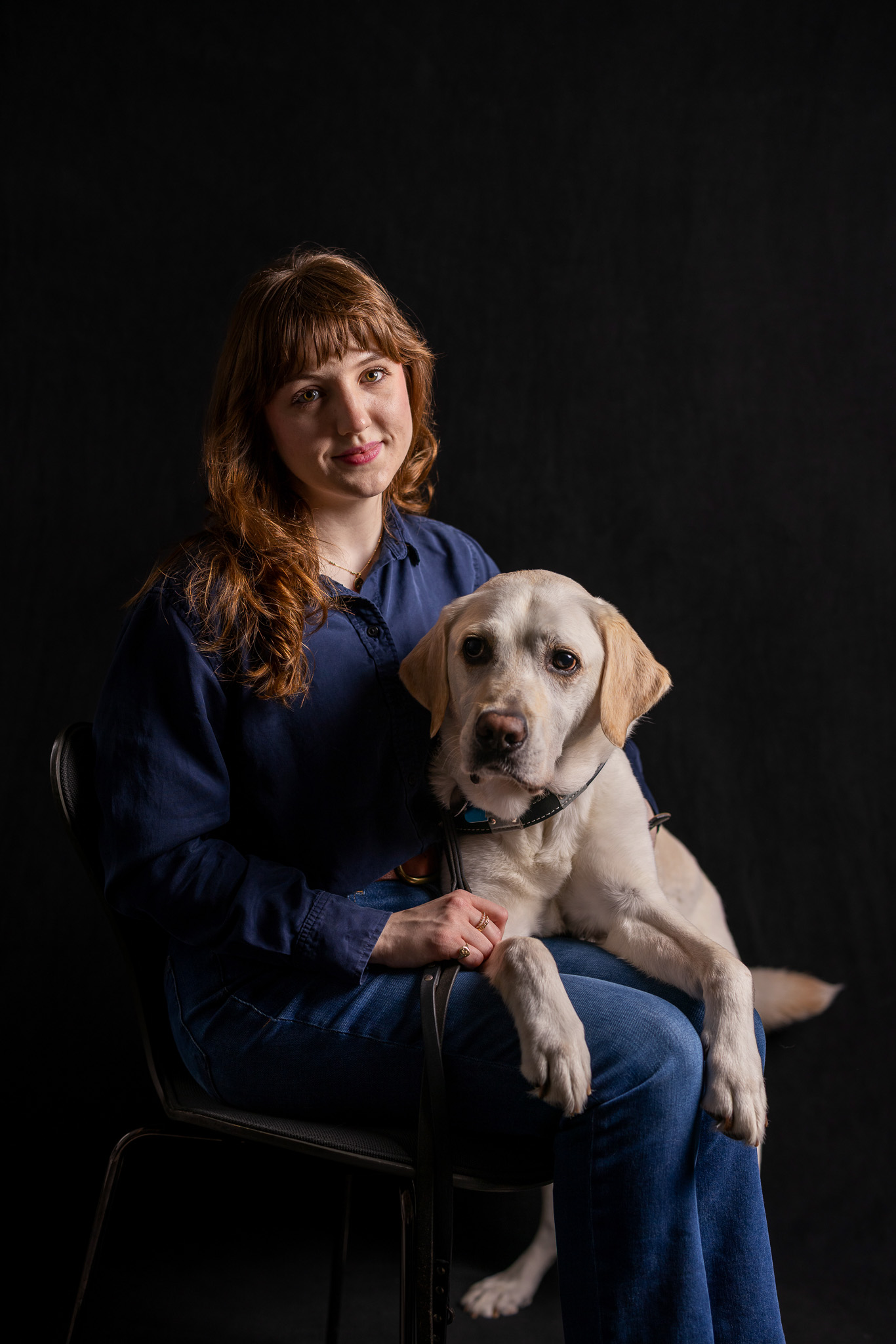 A young woman with brown hair and bangs sits in a chair with a yellow lab on her lap. The woman looks straight ahead with half of her face illuminated by light and the other half in shadow. The background behind her is a dark black.